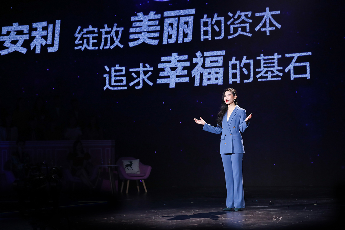 活动现场，身着蓝色西装的女嘉宾站在聚光灯下的舞台上，面带笑意舒展双手正向观众做分享。她身后的星幕大屏显示“安利 绽放美丽的资本 追求幸福的基石”字样，现场氛围积极向上，充分传递安利正向的品牌价值导向。