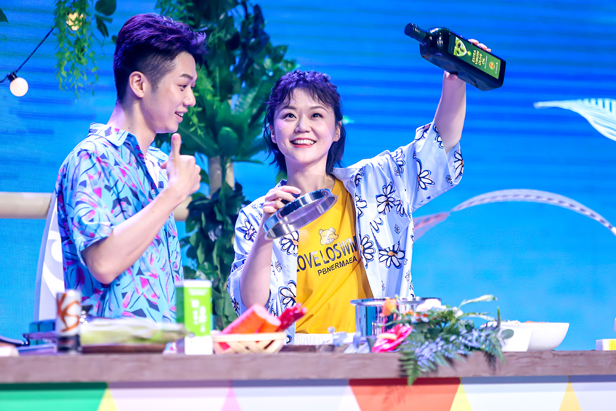 在安利品牌活动的美食互动环节中，女嘉宾高举印有“EXTRA VIRGIN OLIVE OIL”字样的橄榄油产品展示，身旁男嘉宾微笑点赞，台面摆放着新鲜食材与烹饪器具，现场氛围轻松明快，充分传递安利健康优质生活的品牌理念。