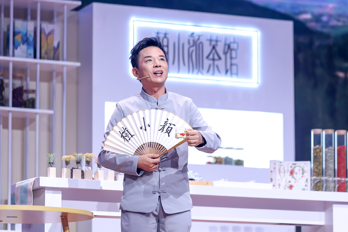 活动背景清晰标注“植小颜茶馆”字样，主讲人身着浅灰色中式服饰，手持印有“植小颜”字样的折扇，端着盛有养生饮品的玻璃杯，现场布置清新雅致，氛围轻松明快，正向观众传递草本健康养生理念，契合安利健康生活的品牌主张。