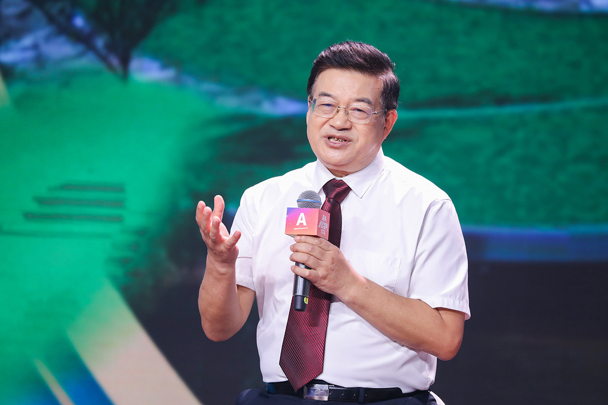 活动现场，特邀嘉宾身着白衬衫搭配酒红条纹领带，手持印有“A”标识的安利活动专属麦克风，站在舞台上配合手势生动分享观点，背景为绿意盎然的大屏，整体氛围专业正向，充分展现活动的务实专业特质。