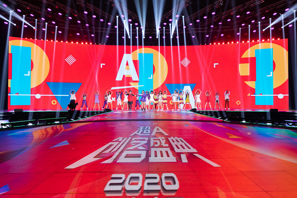 本次是2020安利超A创客盛典活动现场，舞台地面印有“超A创客盛典”“2020”字样，背景大屏搭配醒目“A”字视觉设计，炫彩灯光交汇，一众表演者整齐站在舞台上，姿态昂扬充满活力，现场氛围热烈蓬勃，尽显安利活动的昂扬风貌。