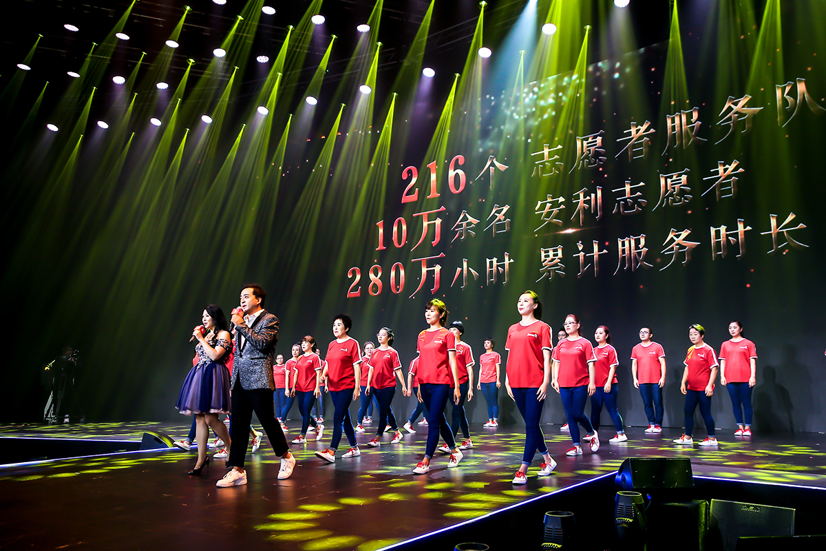 现场灯光绚烂，背景大屏标注着216个志愿者服务队、10万余名安利志愿者、280万小时累计服务时长的公益数据。两名演唱者持麦登台，身着统一红色上衣的安利志愿者整齐列队，整体氛围昂扬热烈，尽显安利志愿者的积极风貌。
