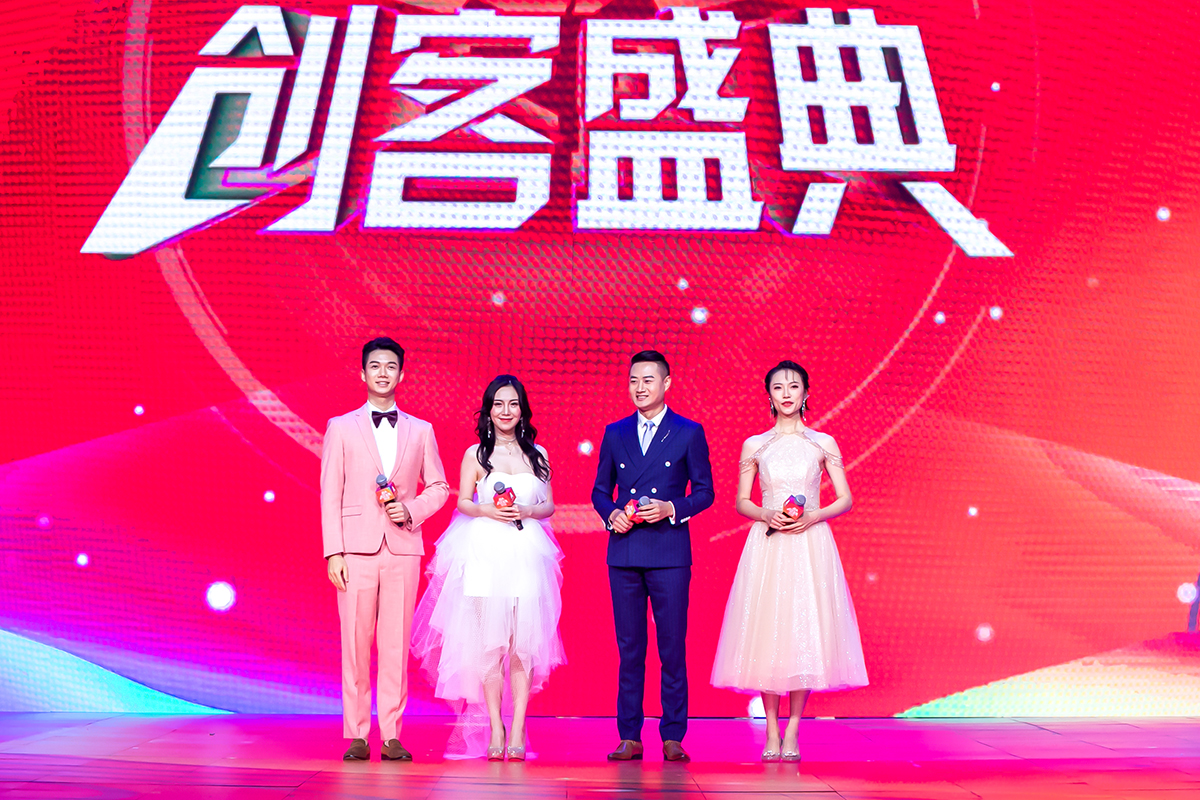 安利创客盛典活动现场，四位着装精致正式的主持人手持话筒站立于舞台上，身后红色大屏醒目展示“创客盛典”字样，现场灯光绚烂、氛围热烈隆重，充分彰显活动的盛大规格与积极向上的品牌风貌。