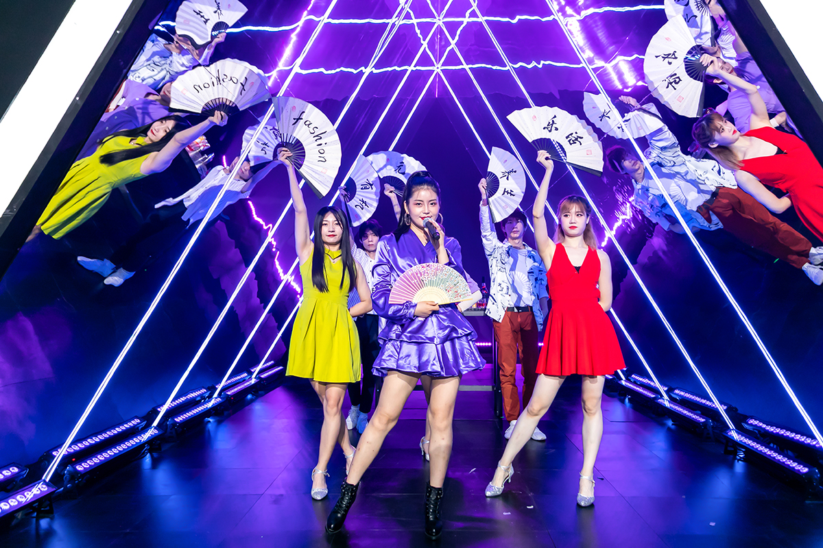 炫紫霓虹灯光打造的潮流舞台上，表演者们手持印有“fashion”“乐活”“养生”等字样的折扇，中心演唱者持麦亮眼出镜，全员状态饱满活力十足，现场氛围热烈鲜活，尽显安利品牌活动的年轻创意与蓬勃朝气。