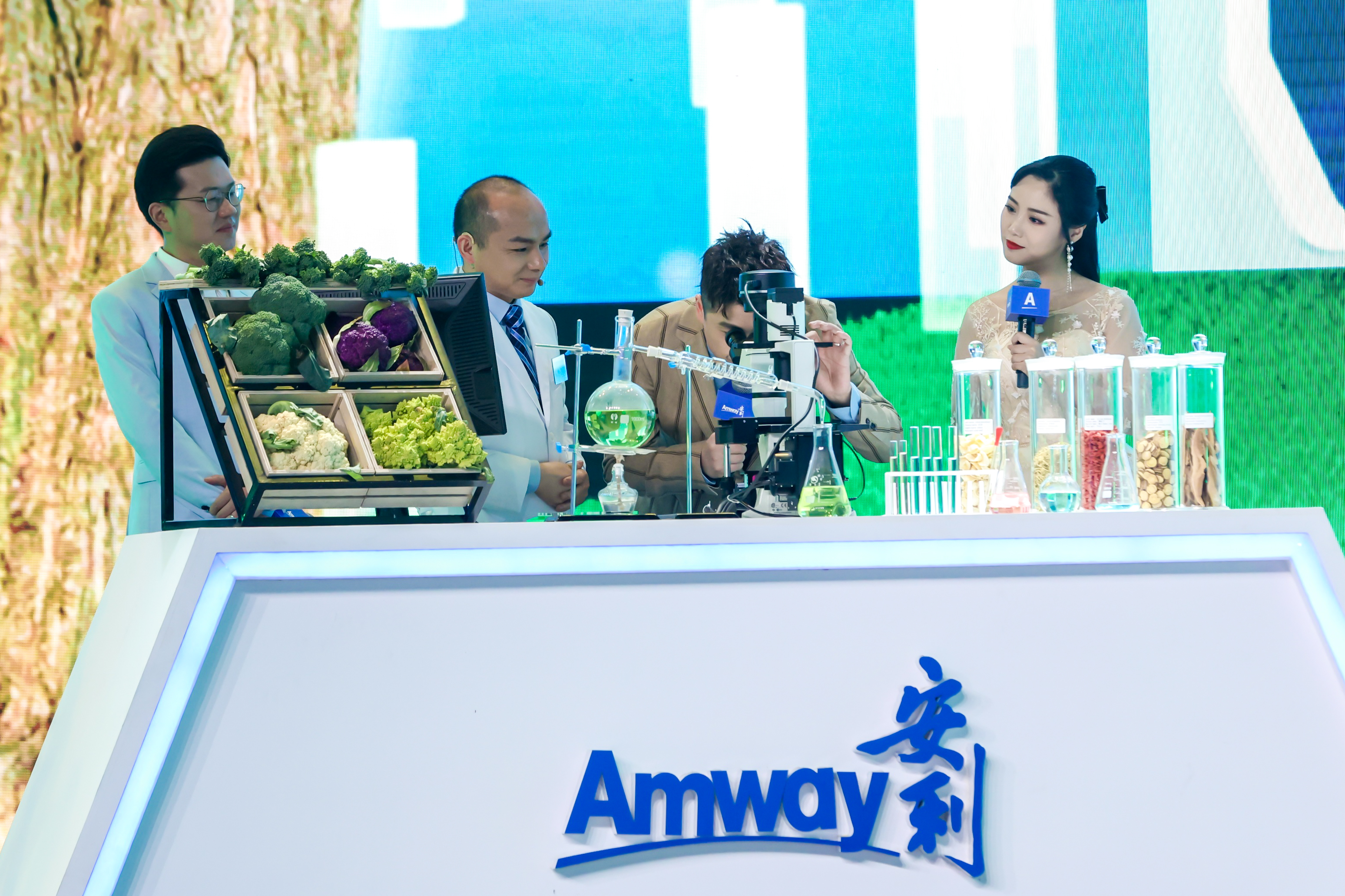 实验台印有“Amway 安利”标识，台上摆放着新鲜蔬果、实验器皿、原料样本，一名人员正操作显微镜观测，旁侧有科研人员、持印有“A”标识话筒的主持人，氛围专业严谨，直观展现安利在营养健康领域的研发实力。