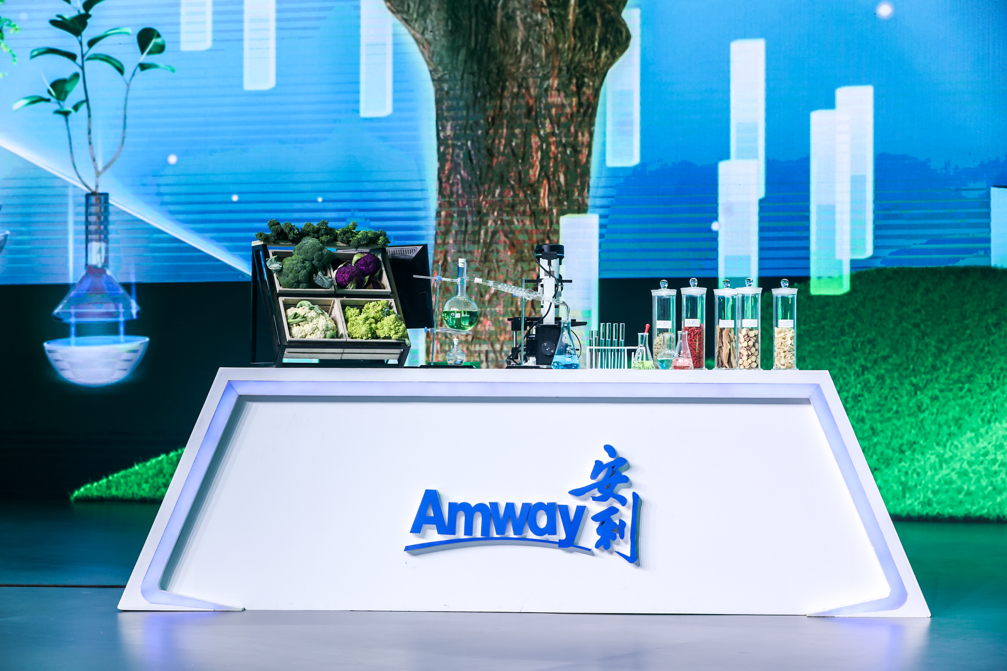 展示台正面印有核心标识“Amway 安利”，台面上摆放着新鲜果蔬、专业实验器具、各类植物原料样本，背景是融合自然与科技感的蓝色大屏，整体氛围专业清新，传递出安利以科技赋能天然营养、深耕优质产品的品牌理念。