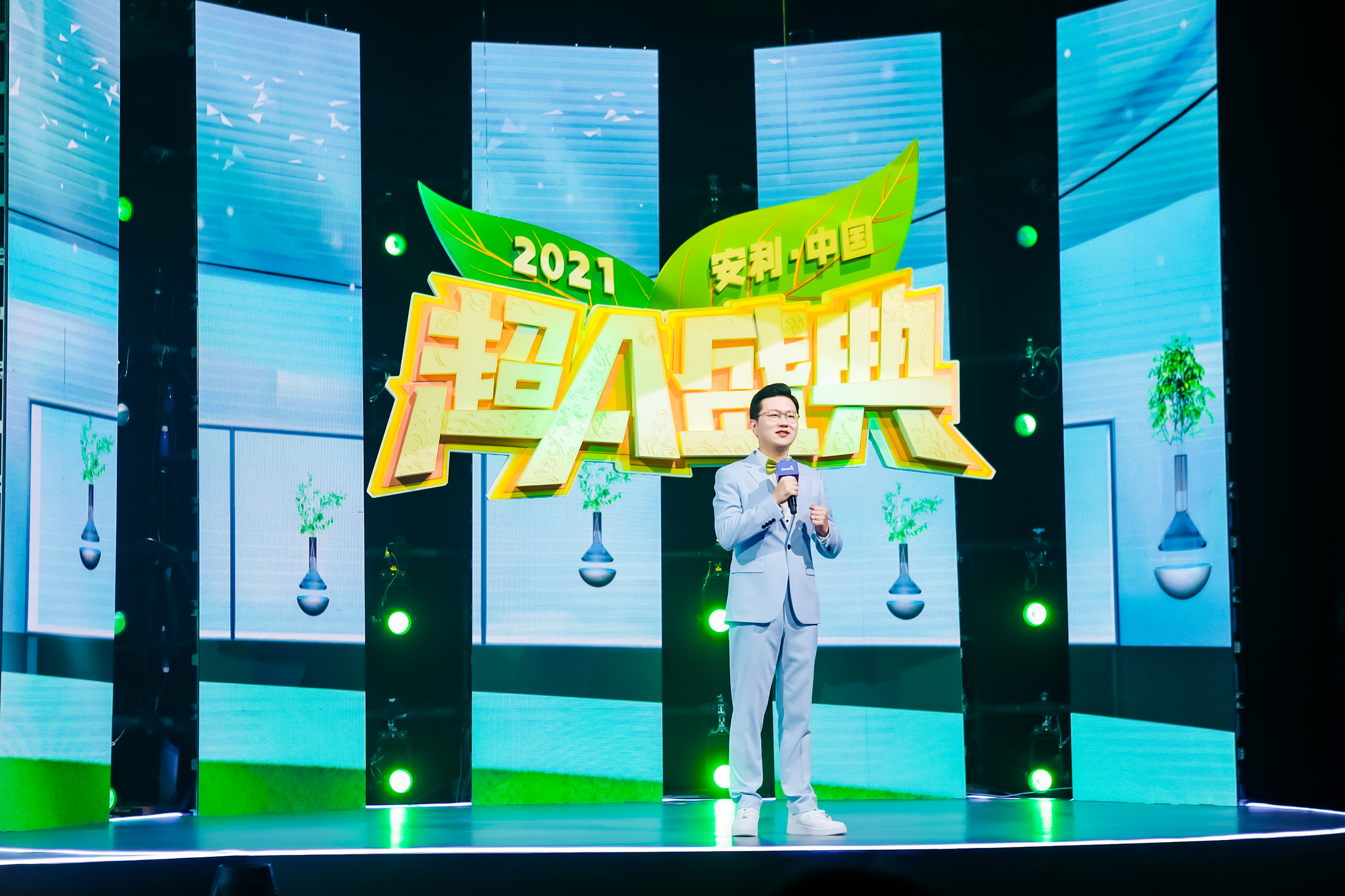 图为2021安利中国超A盛典现场，主持人身着浅蓝西装手持话筒正在舞台上发言，身后醒目设置“2021 安利·中国 超A盛典”主题标识，搭配清新风格LED大屏与绿色灯光，整体氛围明快热烈，尽显品牌活动的活力质感。