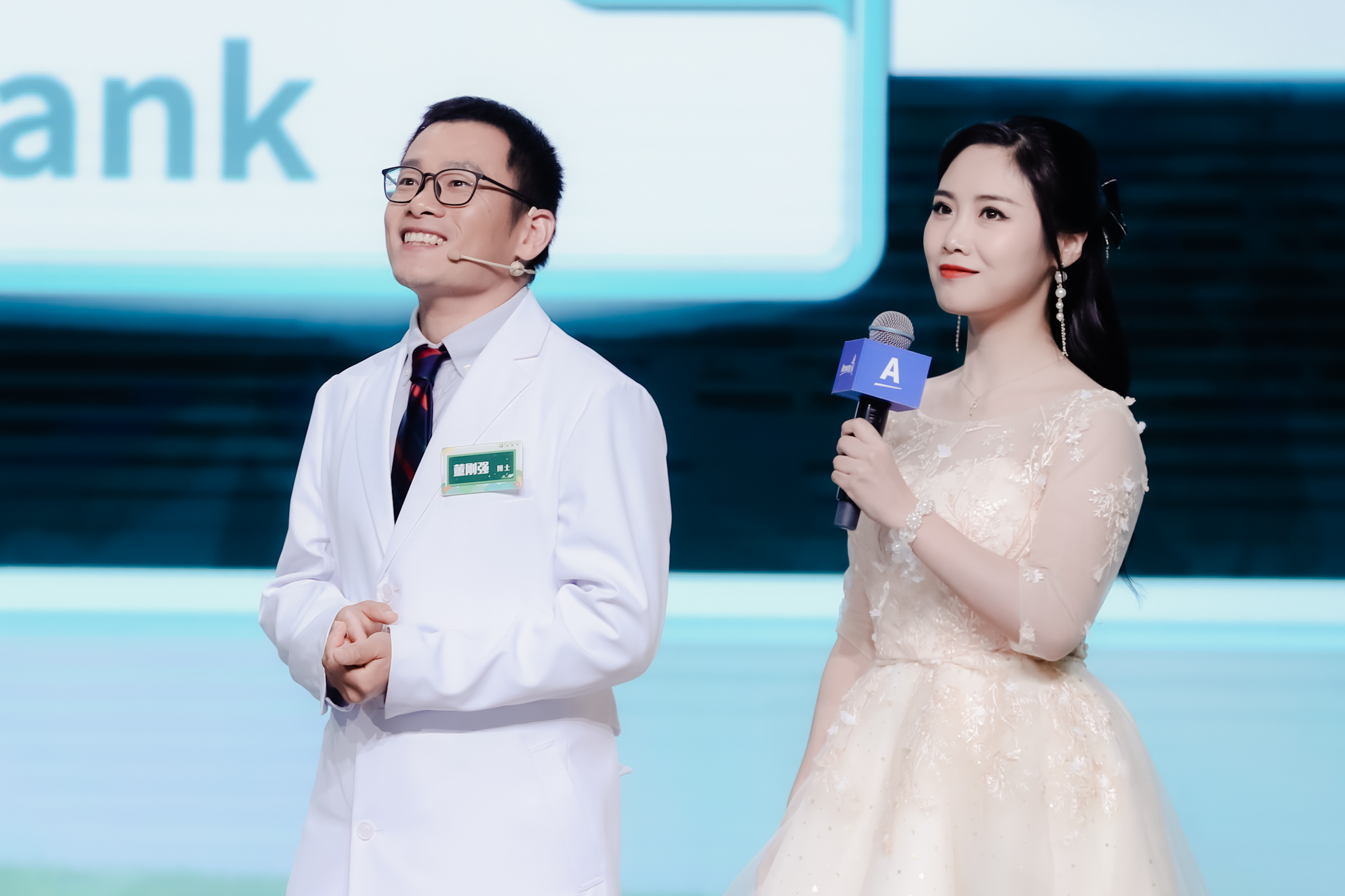 本次安利健康研讨会现场，身着白大褂的董刚强博士与手持印有品牌A标识话筒的女主持人共同站在舞台上，二人状态舒展愉悦，舞台背景清爽明快，整体氛围专业积极，尽显活动的专业质感与活力。
