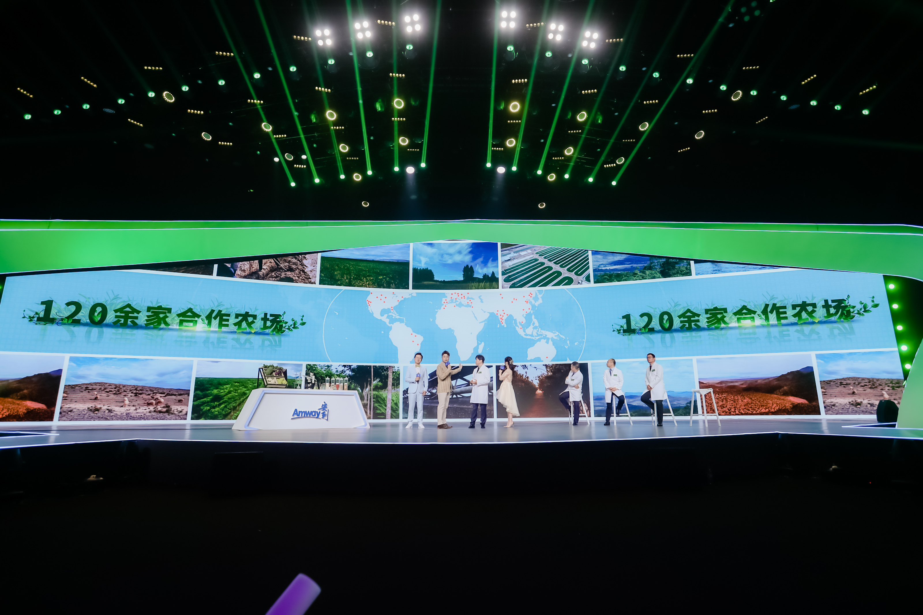 活动舞台背景大屏标注“120余家合作农场”字样，搭配全球农场点位地图与各地农场实景画面，舞台上主持人与多位专家正进行内容分享，台侧设有安利品牌标识台，现场灯光璀璨氛围专业正式，彰显安利原料供应链的硬核实力。