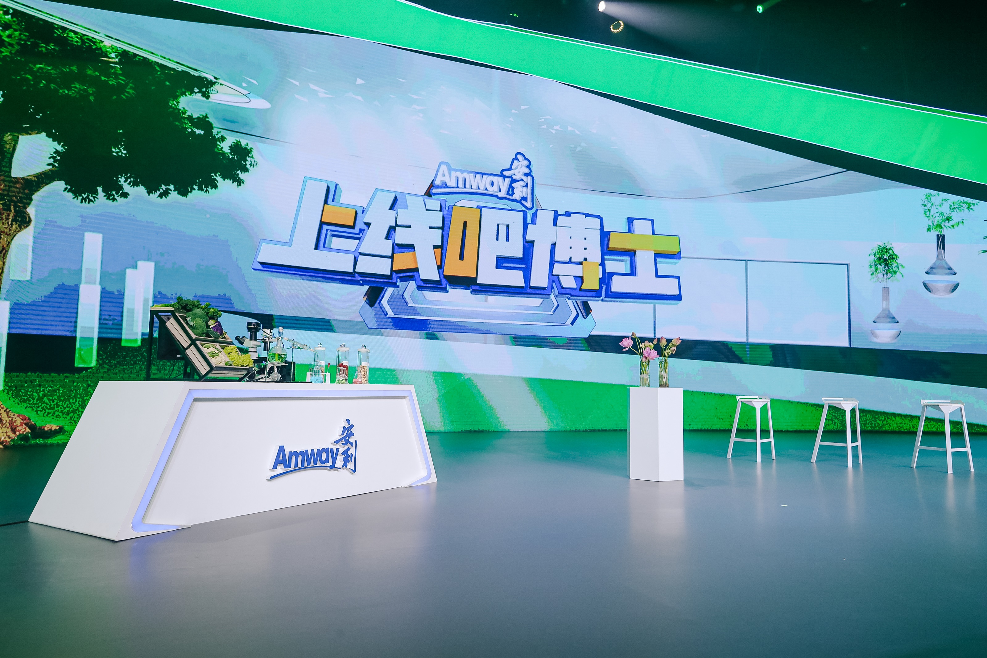 画面为安利《上线吧博士》活动现场，背景大屏醒目展示“Amway安利”与“上线吧博士”主题字样，舞台设置印有安利标识的白色实验操作台，台上摆放实验器具、天然食材样品，旁侧设有花台与白色高脚凳，整体氛围清新明快，兼具科技感与健康属性。