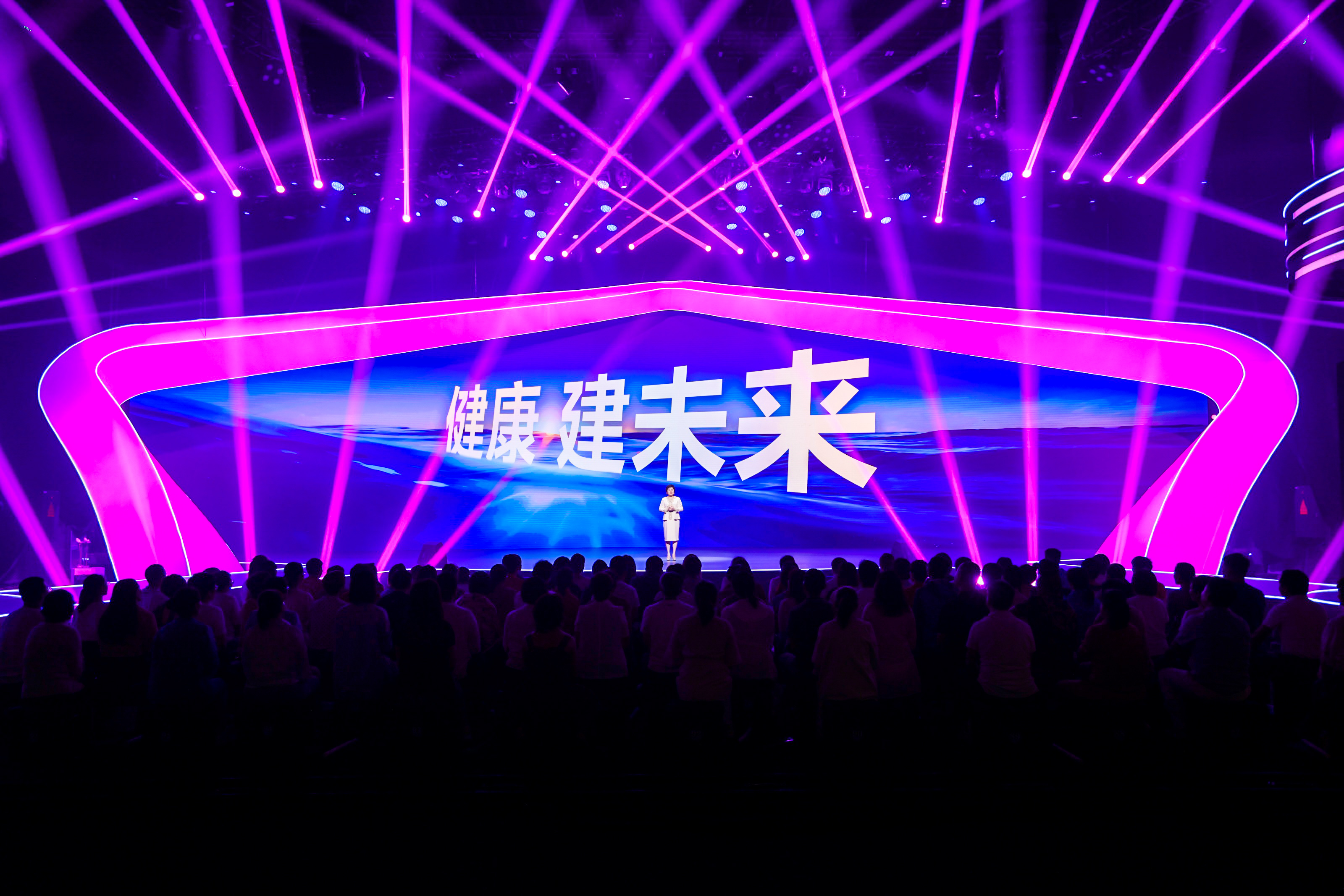 活动现场舞美绚丽，紫粉色灯光交错极具科技感，超大全景主屏幕醒目展示核心主题“健康 建未来”，主讲人站在舞台中央进行分享，台下坐满参会的安利伙伴，现场氛围热烈正式，尽显品牌积极向上、聚焦健康发展的良好风貌。