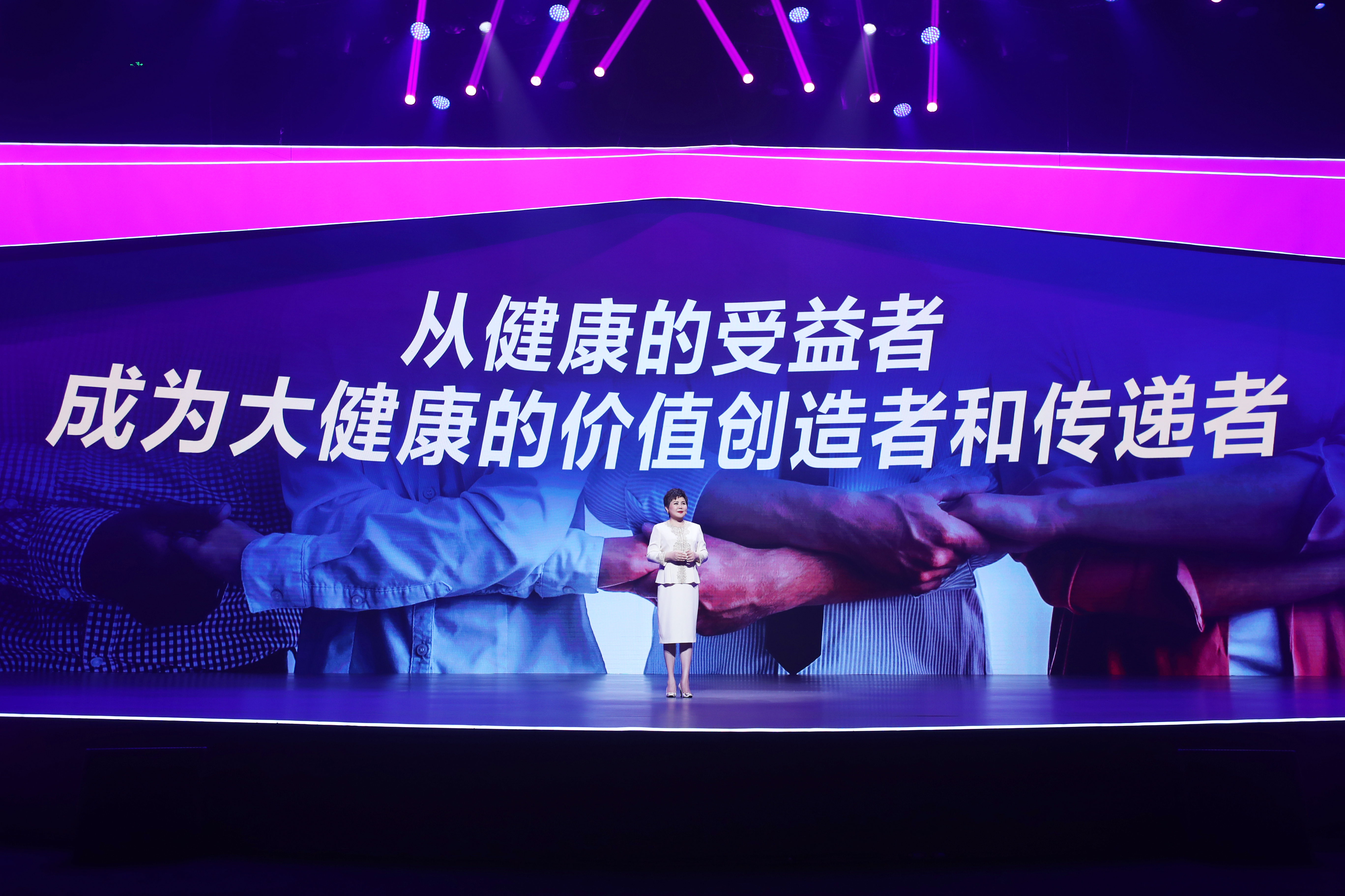 安利品牌活动现场，宣讲人身着白色套装站在灯光绚丽的舞台上，背景大屏醒目展示核心标语“从健康的受益者 成为大健康的价值创造者和传递者”，搭配携手互助的背景画面，氛围庄重热烈，传递出安利深耕大健康赛道、号召伙伴共同传递健康价值的正向导向。