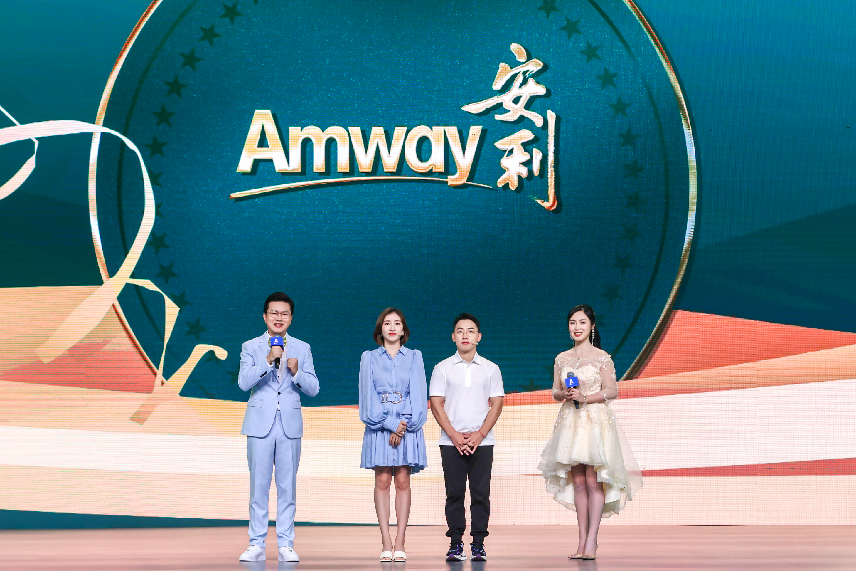 活动现场背景大屏醒目呈现“Amway”与“安利”品牌标识，四位人员站在舞台中央，两位主持人手持印有安利标识的话筒，和两位嘉宾共同登台，现场氛围明亮积极，充分展现出安利品牌活动正式且充满活力的风貌。