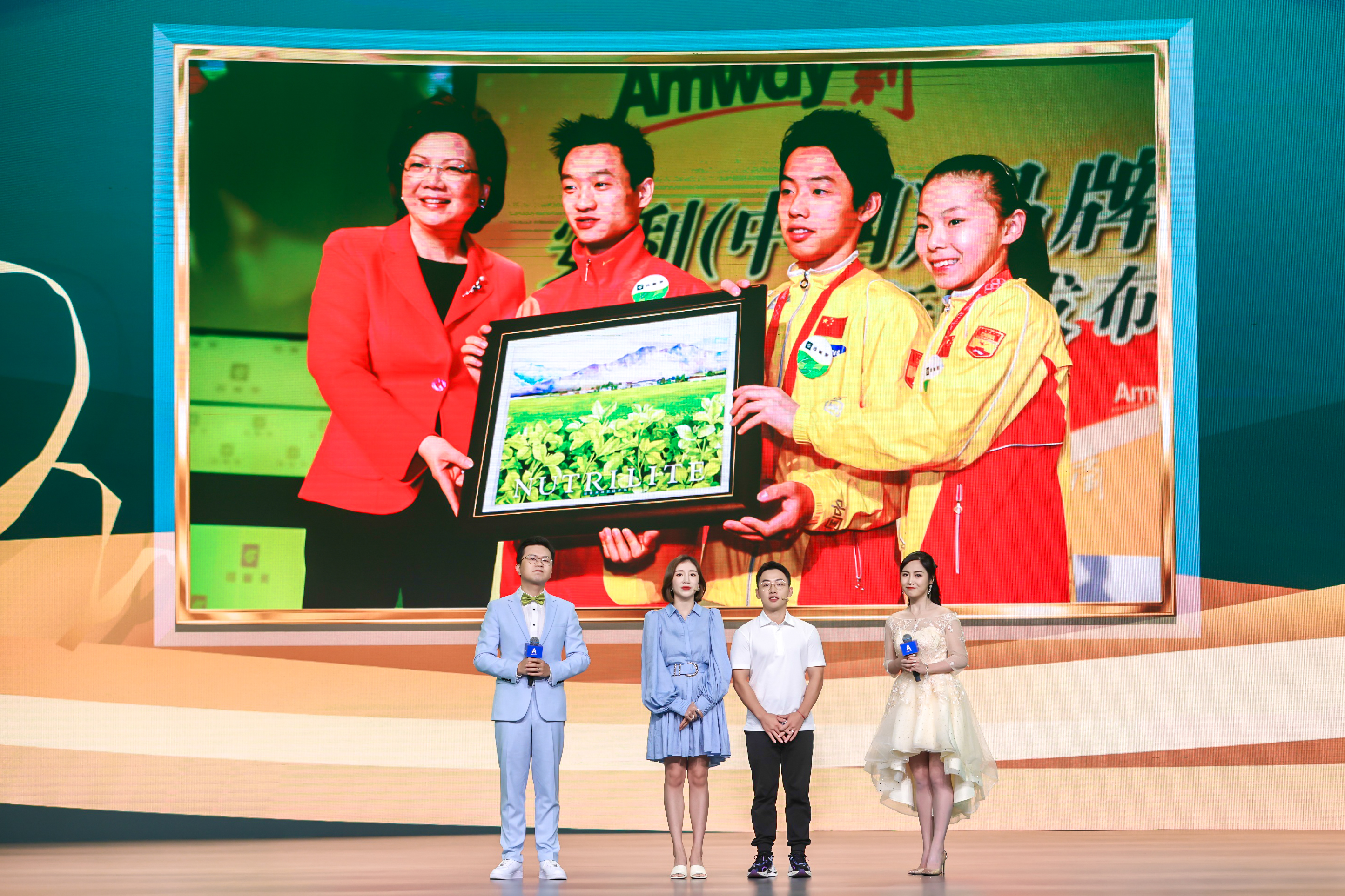本次安利品牌活动现场，四位主持人及嘉宾站立于舞台之上，背景大屏展示着印有“Amway”“NUTRILITE”标识的过往品牌活动合影，记录了安利与运动员代表互赠纪念礼的珍贵时刻，现场氛围正式积极，尽显品牌正向活力。