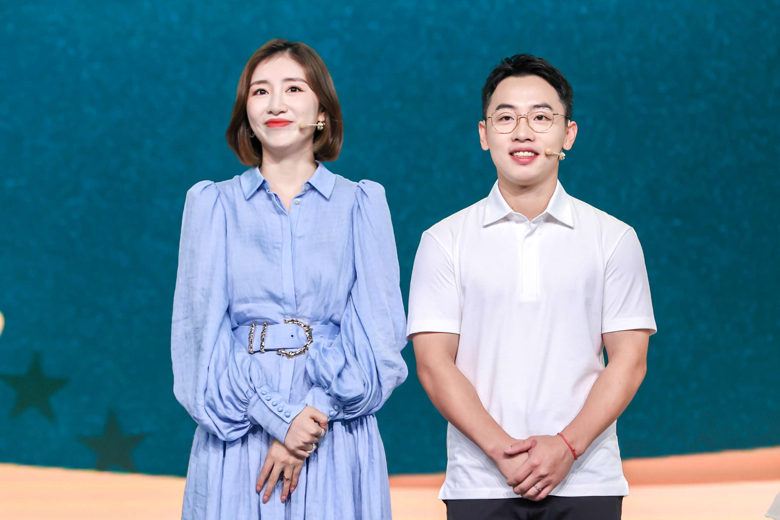 安利品牌活动现场，两位佩戴收音耳麦的主持人登台亮相，女士身着淡蓝色收腰连衣裙，男士穿着简约白色POLO衫，二人面带明朗笑容立于青蓝色带星饰的舞台背景前，状态饱满亲和，现场氛围明快正向，尽显活动的专业活力调性。