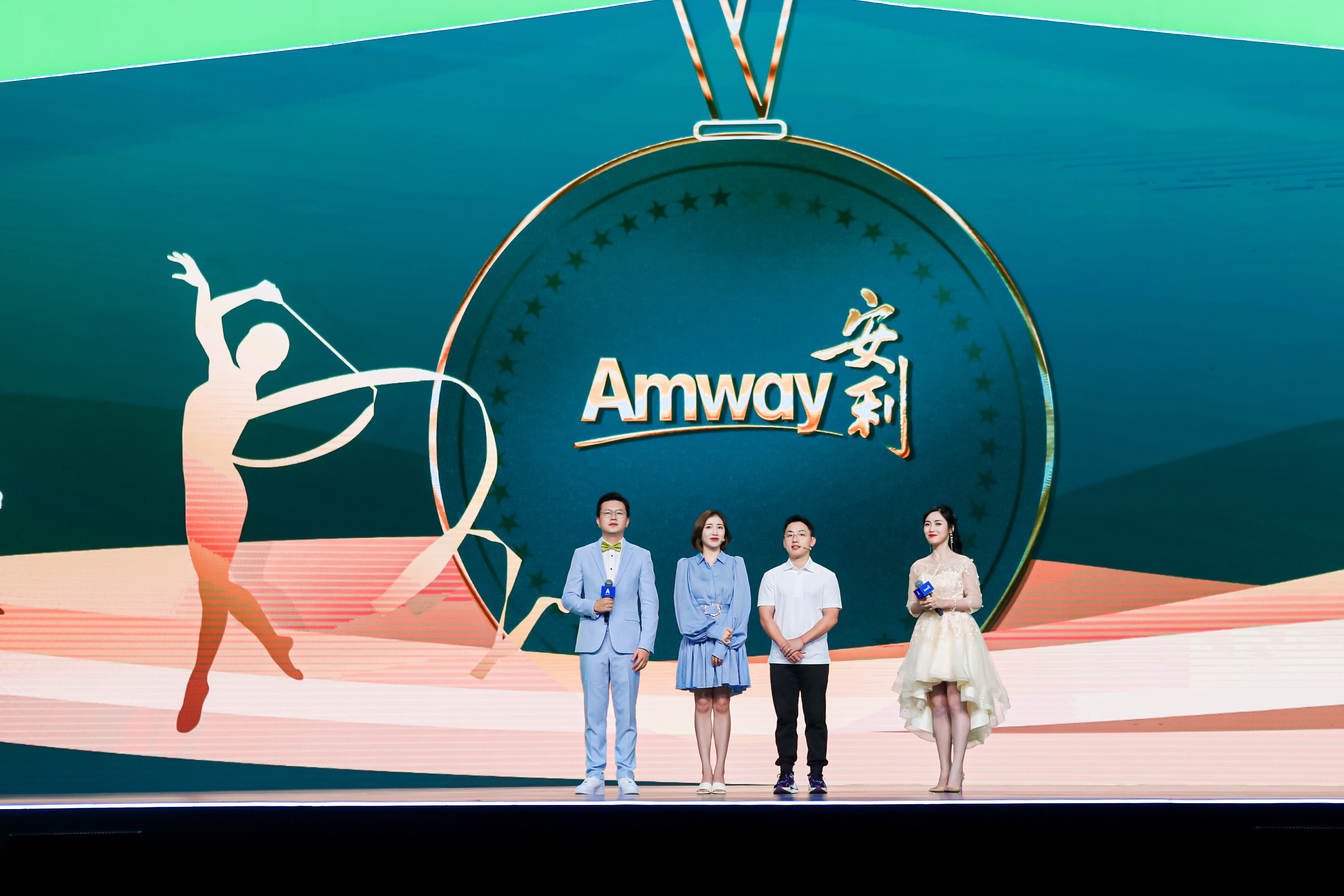 本次安利品牌活动现场，背景大屏呈现带有“Amway 安利”字样的奖牌造型视觉与艺术体操运动剪影，四位人员站在舞台之上，其中两位主持人手持话筒，整体氛围明快昂扬，尽显活动正式感与积极向上的品牌风貌。