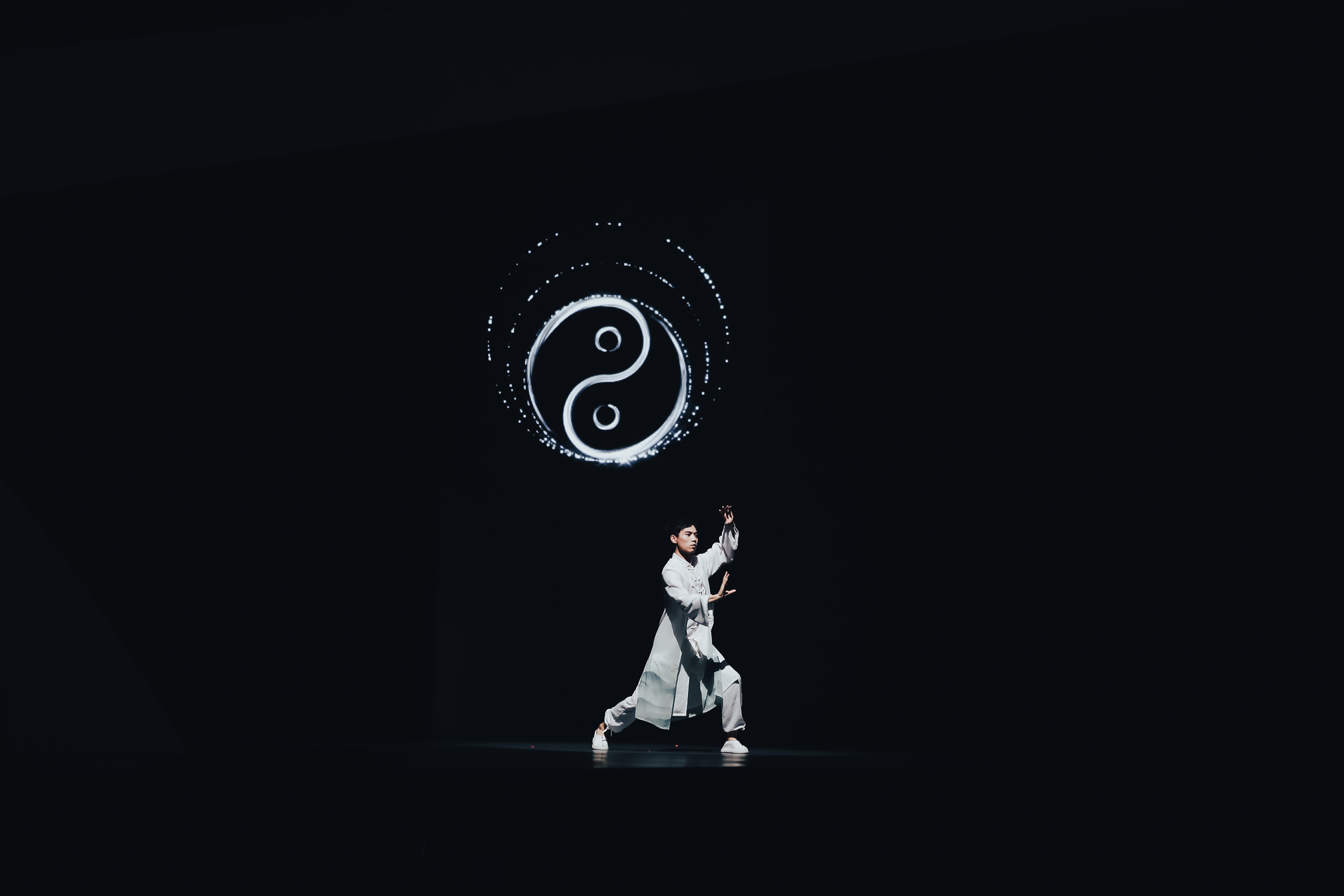 安利品牌活动现场，身着浅白中式练功服的表演者正在舞台上展演太极，身后悬浮着发光的太极阴阳符号，暗调舞台衬得表演更具沉静大气的东方气韵，传递出平衡健康的养生理念，契合安利倡导健康生活的品牌调性。