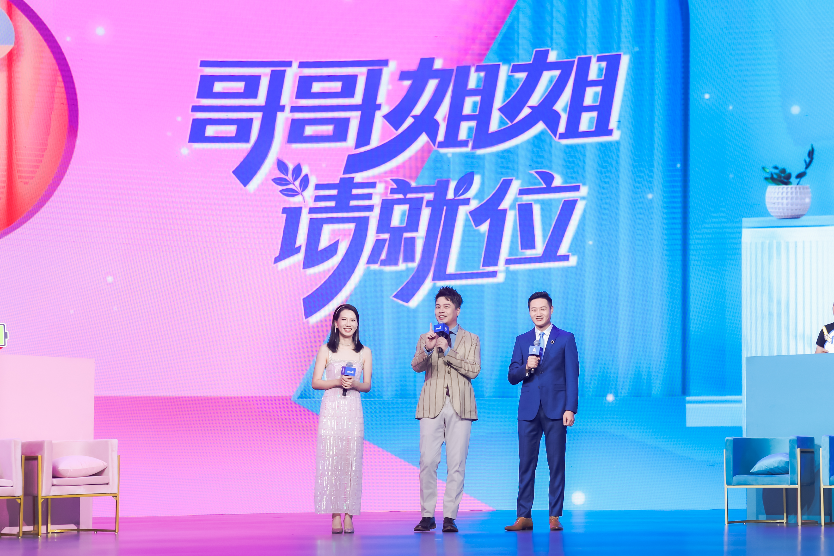 安利活动现场背景大屏标注核心文字“哥哥姐姐请就位”，三位主持人手持话筒站在粉蓝渐变的活力舞台上，状态饱满笑容亲和，现场氛围明快活泼，充分展现出安利品牌活动积极鲜活的风格，传递出轻松正向的活动氛围。