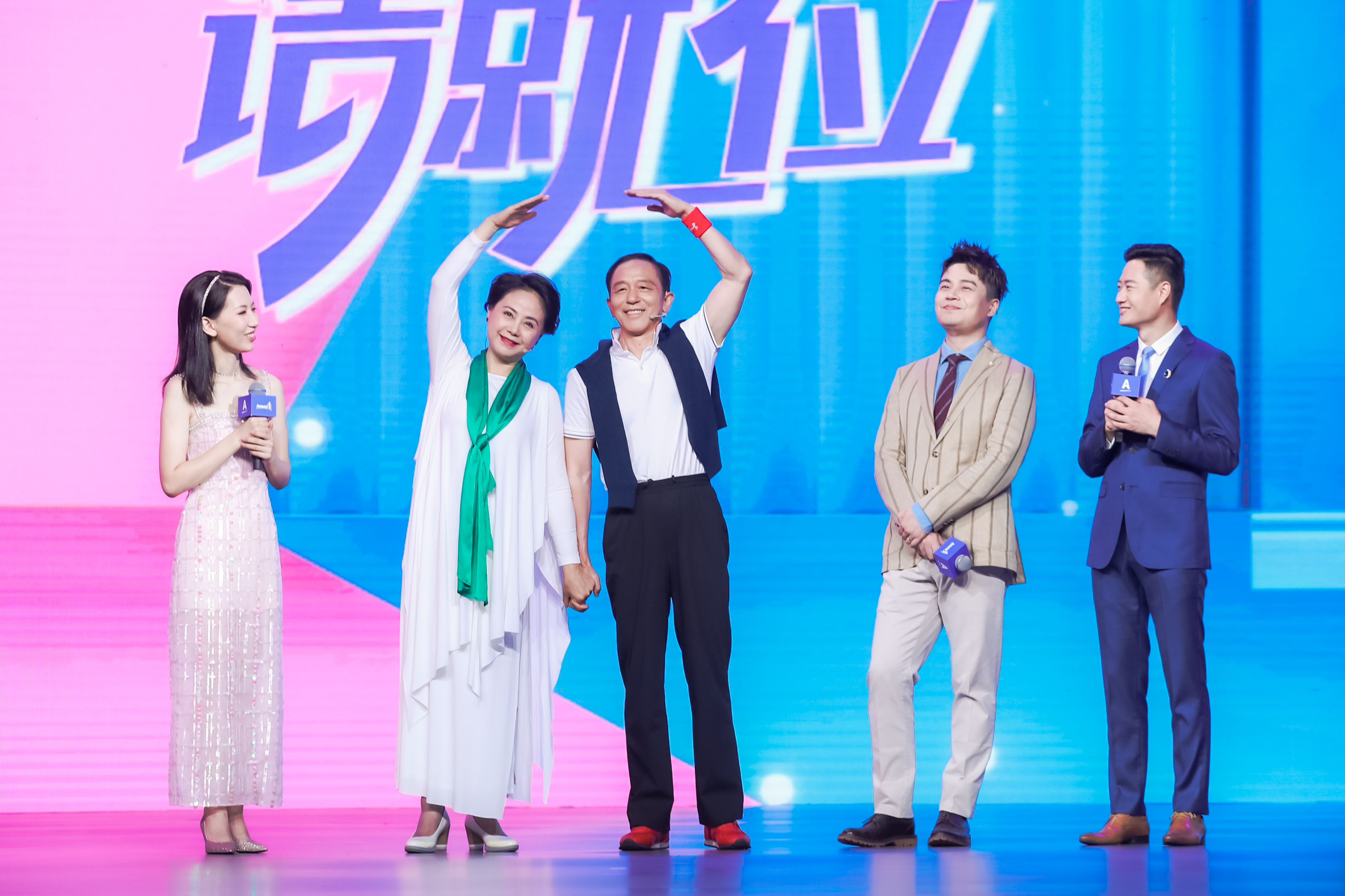 安利品牌活动现场，粉蓝渐变舞台背景显示核心文字“请就位”，五位嘉宾齐聚舞台，一对伙伴牵手共同比出爱心手势，笑容温暖明朗，其余三位嘉宾含笑在侧，现场氛围轻松热烈，尽显安利伙伴融洽向上的良好风貌。