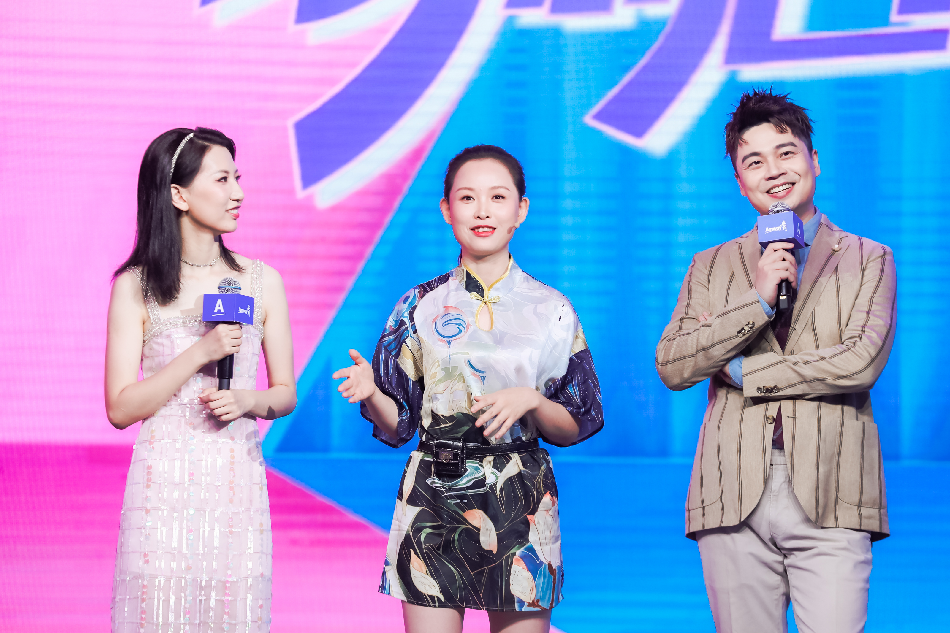 活动现场以粉蓝渐变炫彩大屏为背景，三位人员站在舞台之上，两人手持印有“Amway”标识的话筒，中间身着新中式服饰的嘉宾正抬手交流发言，现场氛围轻松热烈、积极鲜活，彰显安利品牌活动的活力风貌。