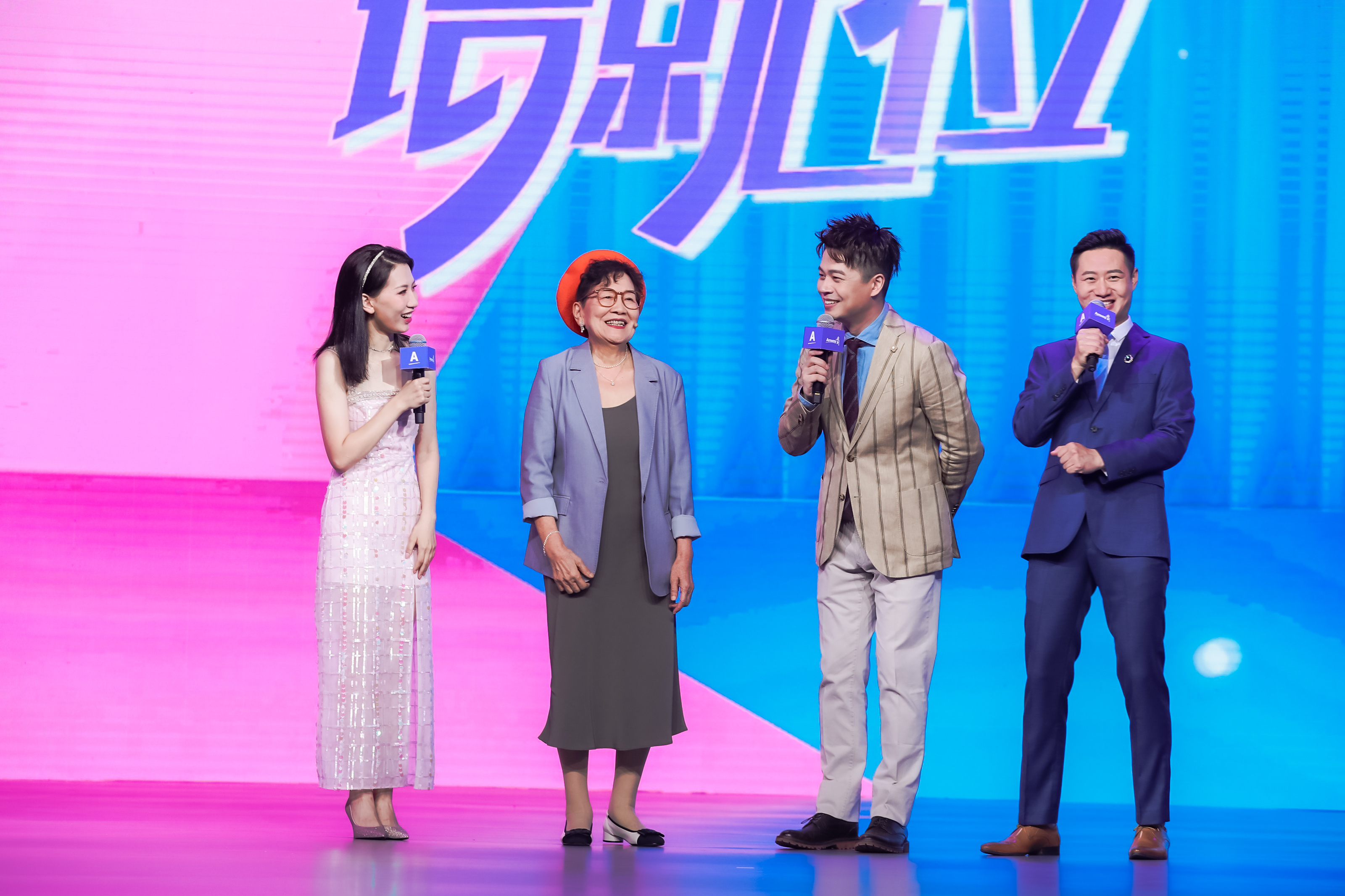 安利品牌活动现场，粉蓝渐变背景大屏显示“场就位”字样，三位手持印有安利标识麦克风的主持人，与受邀到场的年长嘉宾在舞台上轻松交流，众人笑意融融，现场氛围热烈明快，尽显安利活动温暖积极的基调。
