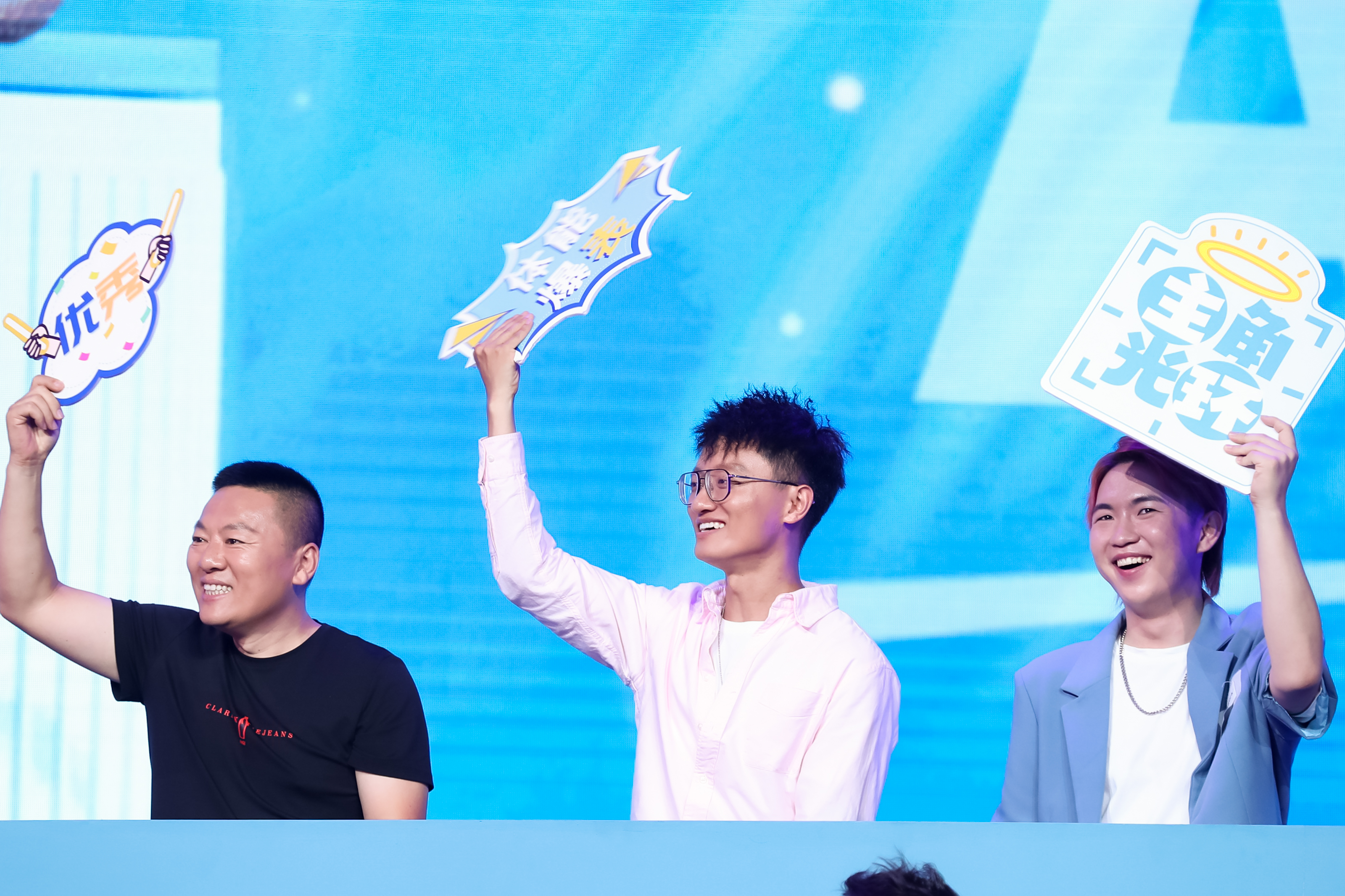 在安利品牌活动现场，三位参会伙伴面带灿烂笑意，分别高举印有“优秀”“体能爆表”“主角光环”字样的趣味手举牌，身后是清爽的蓝色活动大屏，整体氛围轻松热烈，充分展现出参与者昂扬饱满的状态与活动鲜活的互动氛围。