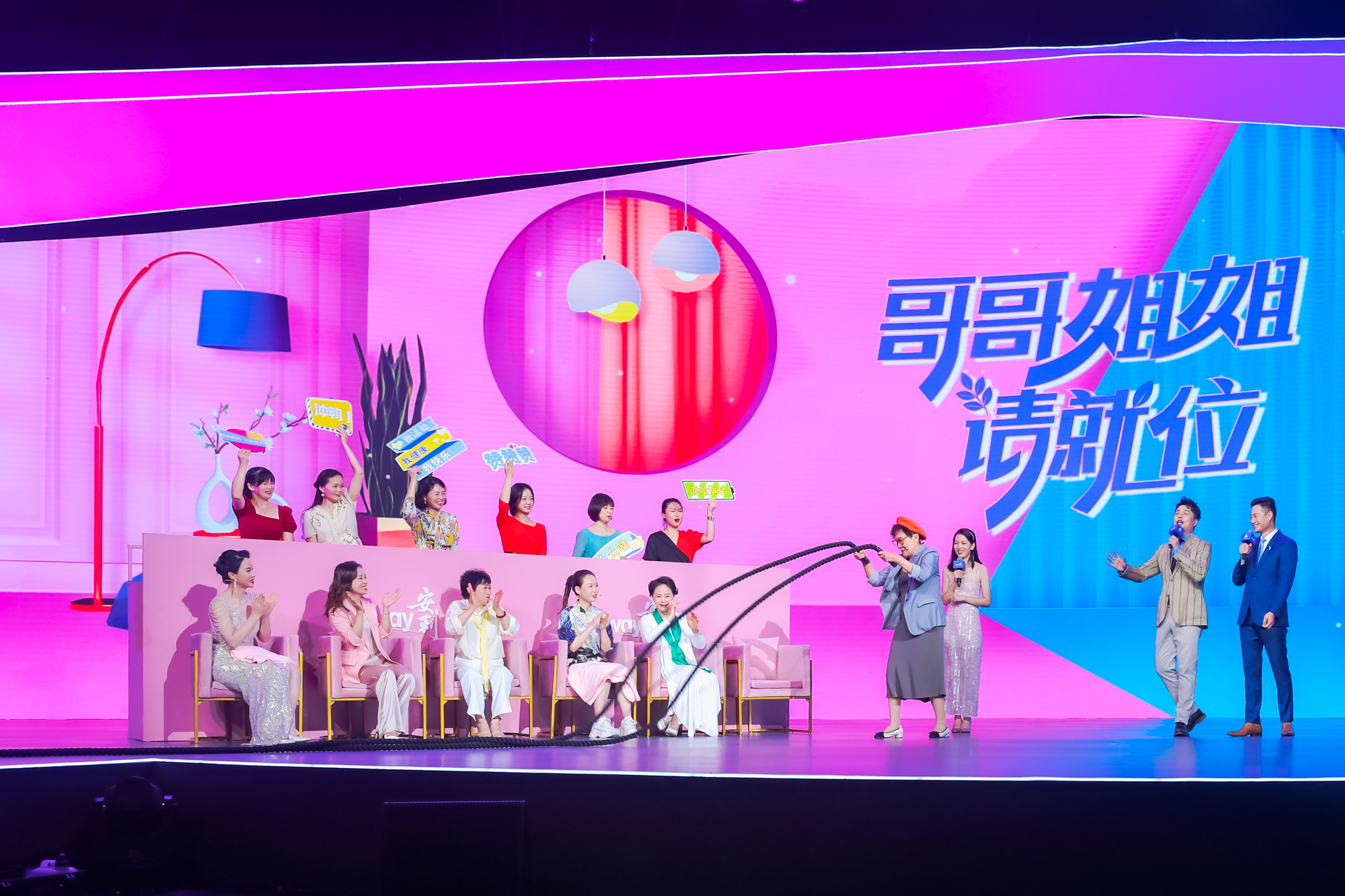 这是安利品牌《哥哥姐姐请就位》互动活动现场，舞台背景醒目展示活动主题字样，台上嘉宾有的手持“冲鸭”“赞赞赞”等应援牌欢呼，有的落座鼓掌助威，参与者正开展趣味互动，主持人在旁引导，现场氛围轻松热烈，尽显安利活动的活力与融洽氛围。