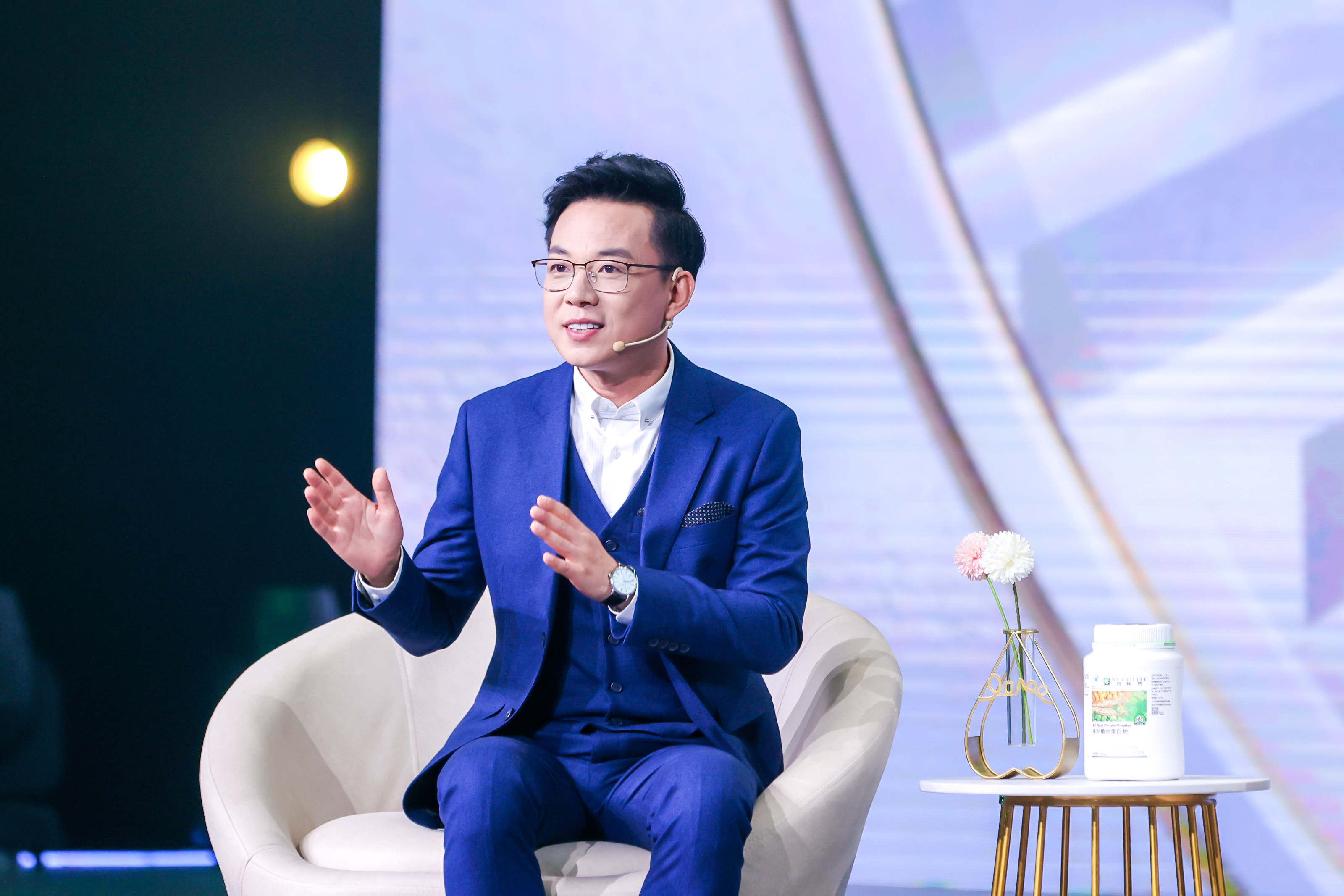 安利研讨会现场，身着蓝色西装的嘉宾佩戴耳麦坐于沙发上，面带笑意抬手交流，氛围专业明快。旁侧边几摆放着纽崔莱多种植物蛋白粉，整体场景积极正向，充分展现出安利品牌专业的健康理念分享氛围。