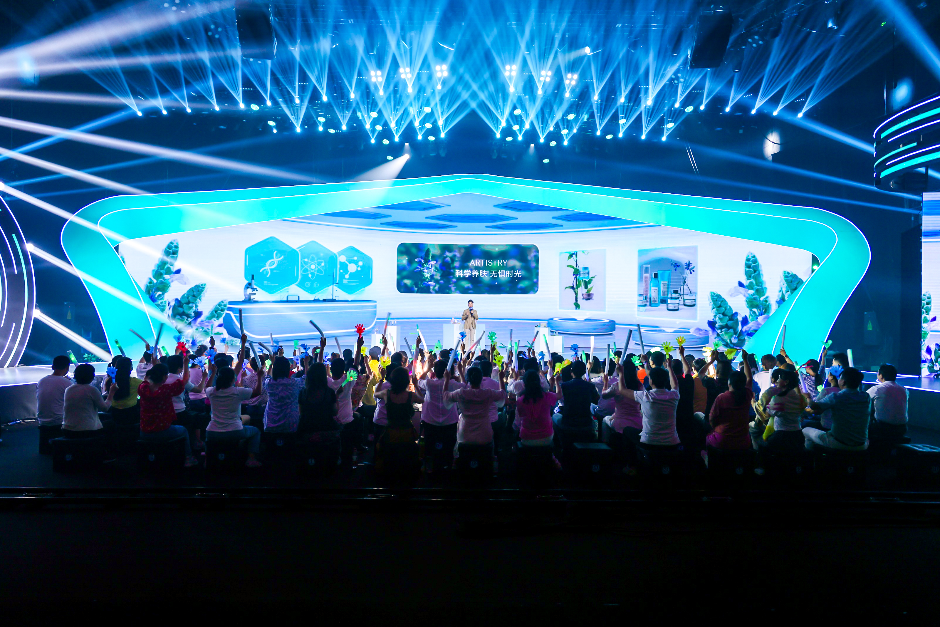 活动舞台采用科技感舞美设计，背景展示核心标语「ARTISTRY 科学养肤 无惧时光」，讲师正站在台上进行分享。台下参会伙伴热情高涨，纷纷举起应援道具呼应，现场灯光绚烂氛围热烈，充分展现安利品牌活动的高参与度与活力。