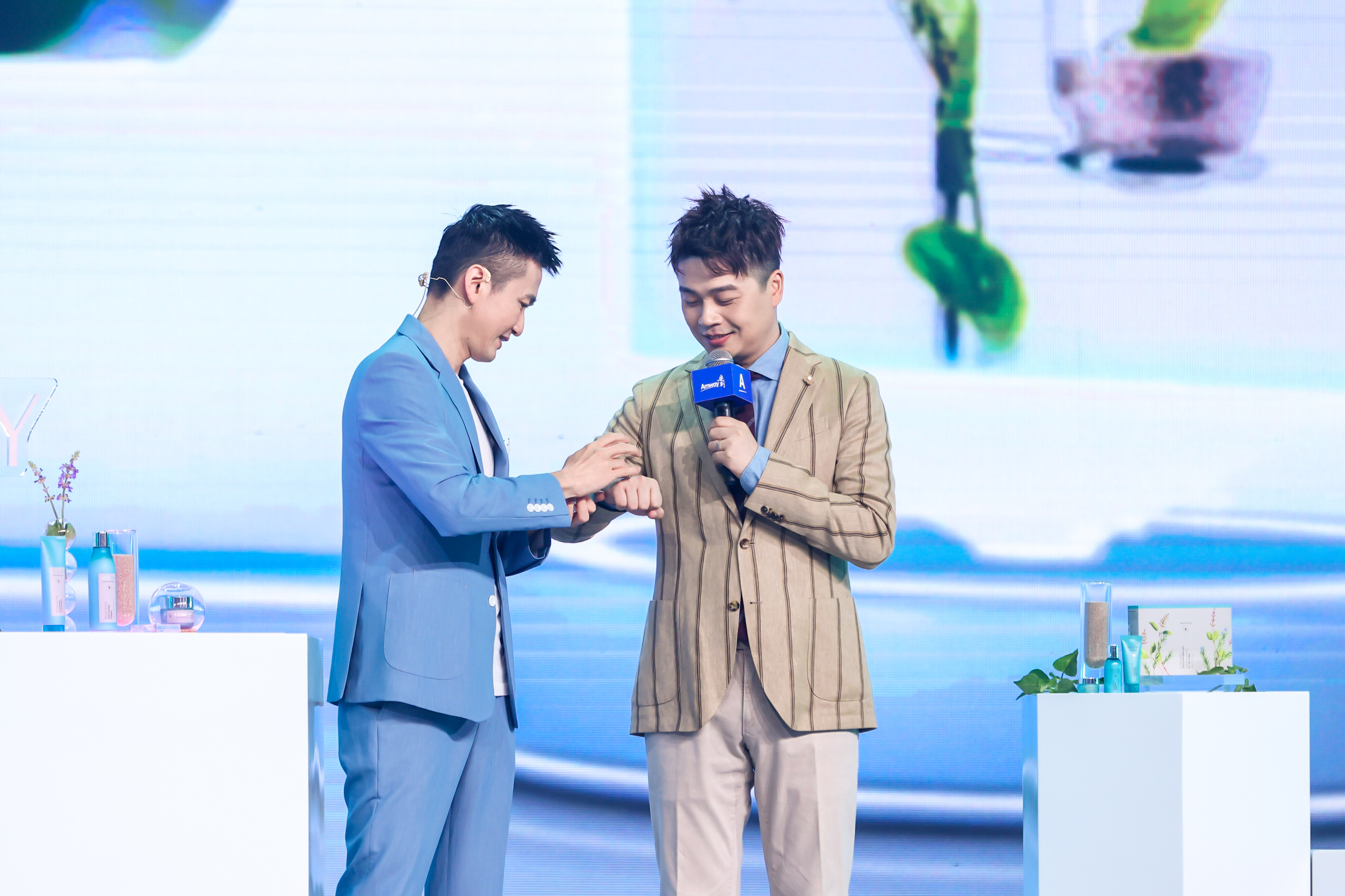 安利活动现场，一位男士手持印有“Amway”标识的话筒，另一位嘉宾正在为他试用护肤产品，舞台两侧展台上陈列着多款安利护肤产品，背景为清新风格大屏，整体氛围轻松专业，直观展现安利产品的使用体验，传递品牌优质护肤理念。