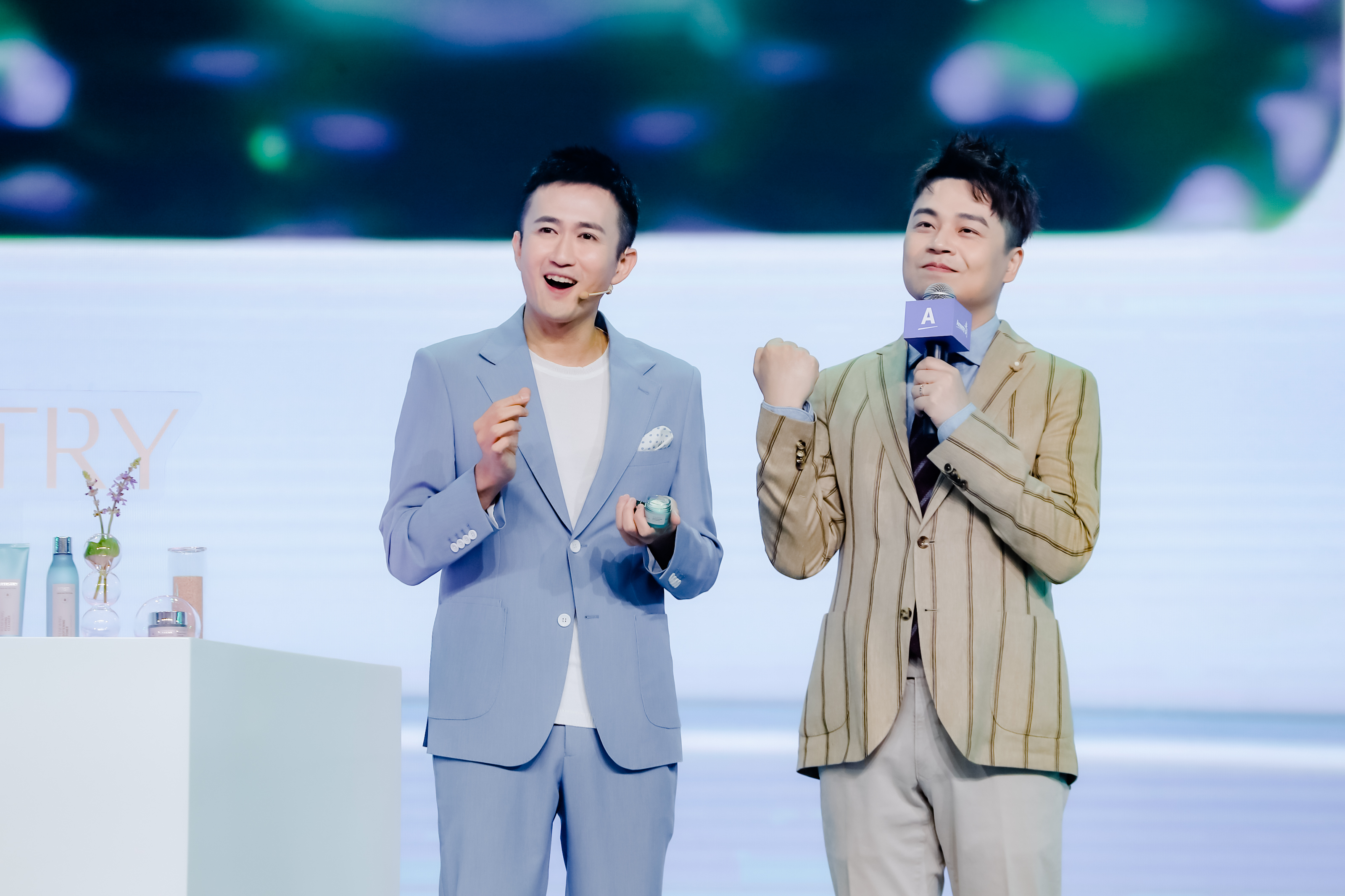 安利活动舞台上，两位嘉宾正在开展互动，一位手持护肤产品进行内容讲解，另一位手持印有“A”标识的品牌话筒积极回应，旁侧展台上陈列着多款安利护肤产品，现场氛围轻松热烈，尽显安利专业积极的品牌调性。