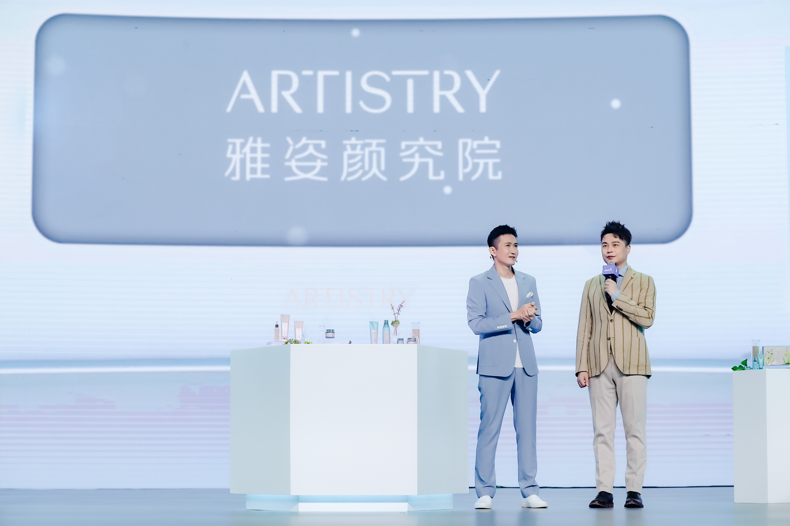 活动现场背景大屏醒目呈现“ARTISTRY 雅姿颜究院”字样，两位嘉宾站在舞台上交流分享，前方白色展台上陈列着多款雅姿护肤产品，整体氛围专业明快，充分传递雅姿品牌的专业护肤理念与鲜活特质。