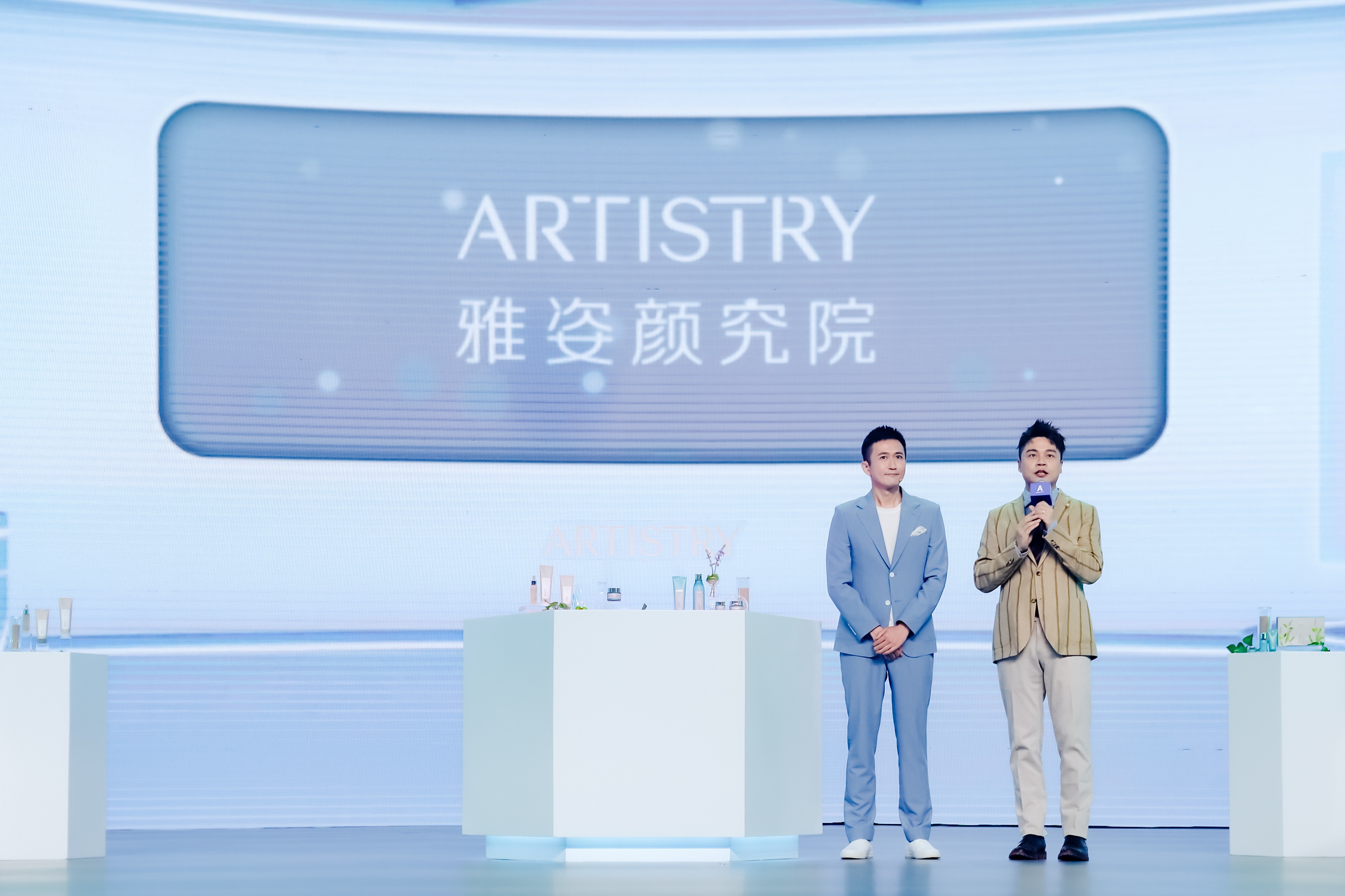 活动现场背景大屏展示“ARTISTRY 雅姿颜究院”核心字样，两位男士立于台前，其中一位手持话筒正在分享讲解，展台上陈列着雅姿系列护肤产品，整体场景明亮清爽，氛围专业积极，充分展现雅姿品牌的专业护肤调性。