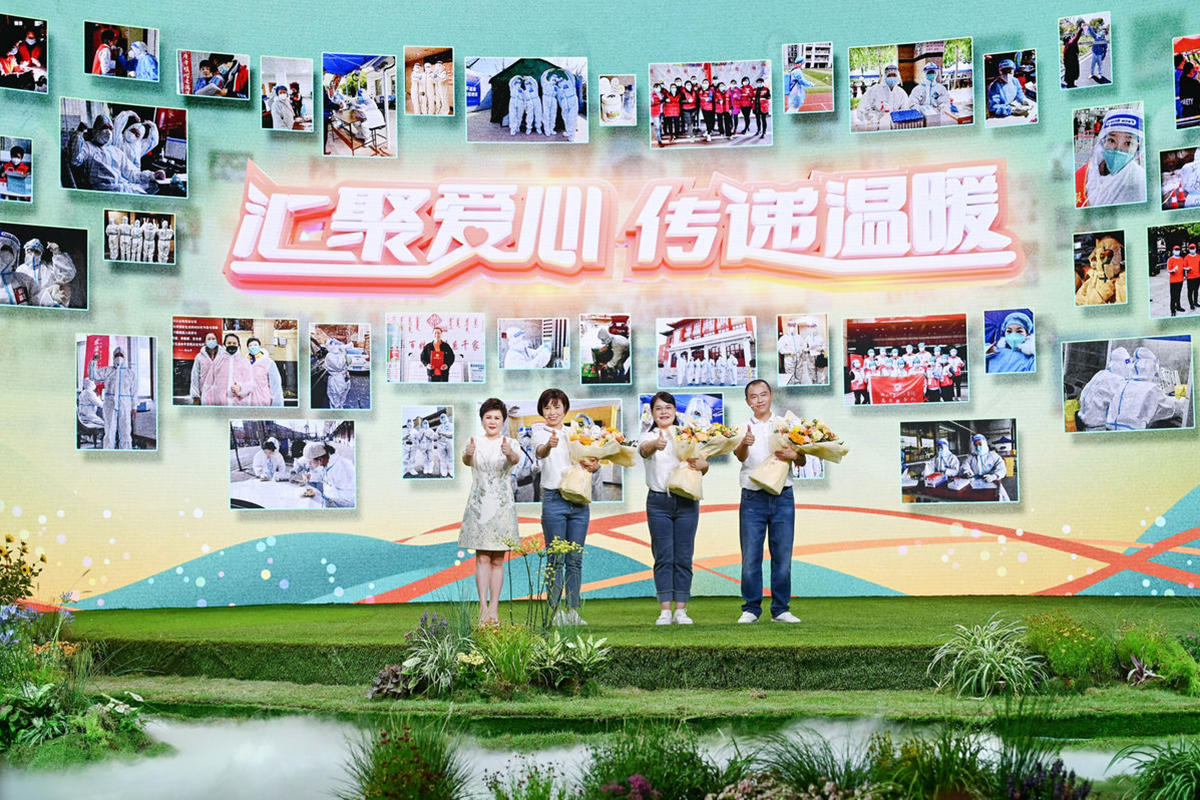 活动背景板核心标语为“汇聚爱心 传递温暖”，板上布满安利抗疫志愿服务纪实照片，四位优秀抗疫志愿者代表手持献花站立于舞台上，一同竖起大拇指，现场氛围温暖昂扬，彰显出安利践行社会责任、传递公益大爱的品牌理念。