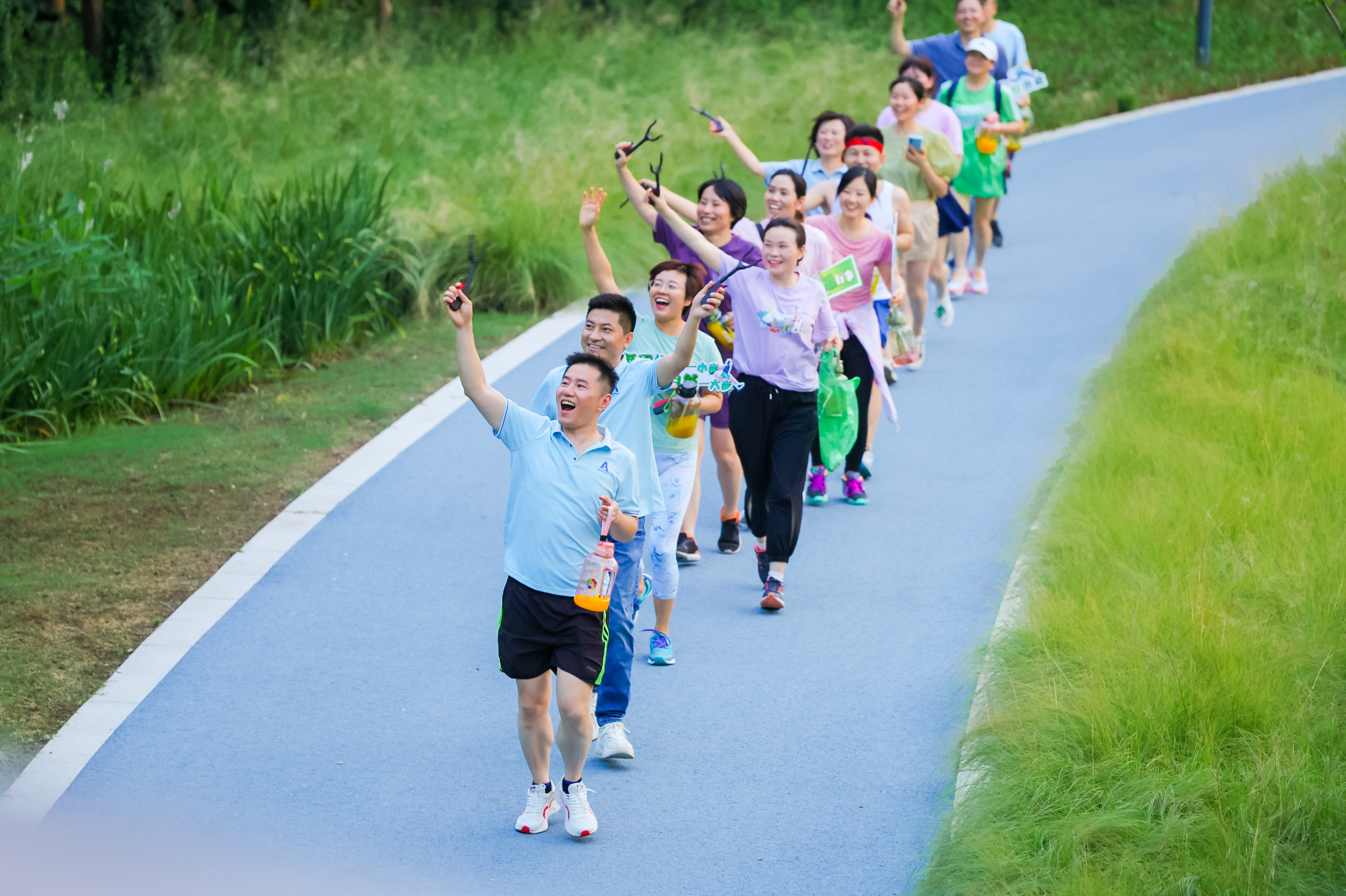 户外生态步道上，参与安利公益捡跑活动的伙伴们列队前行，大家手持垃圾捡拾夹、环保垃圾袋，面带灿烂笑容，情绪饱满高昂，充分展现出安利人热心环保公益、昂扬向上的良好风貌，现场氛围活力十足、充满正能量。