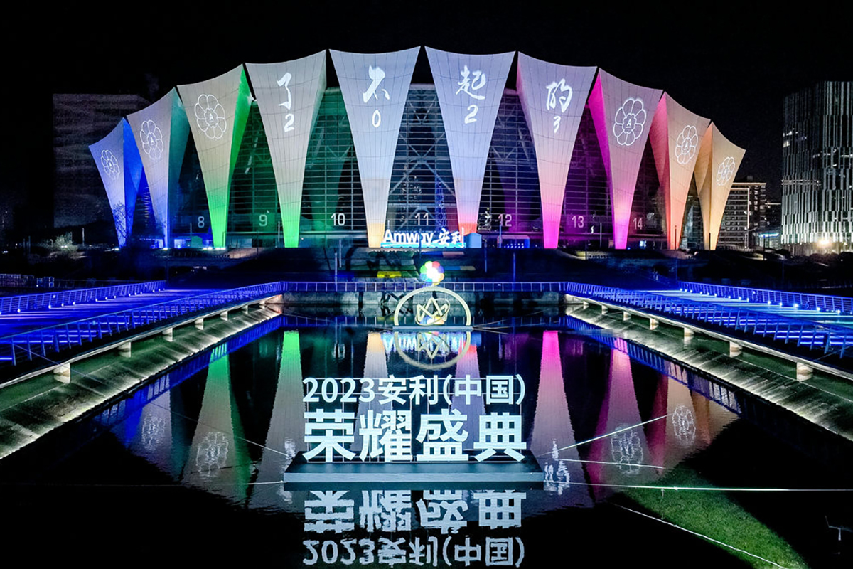 夜幕下，活动场馆被绚丽渐变彩光点亮，外立面设有Amway安利标识，还投影了“了不起的2023”字样。场馆前方水面设置“2023安利（中国）荣耀盛典”发光立体字，灯光与水中倒影交相辉映，整体氛围恢弘璀璨，尽显此次品牌活动的盛大规格。
