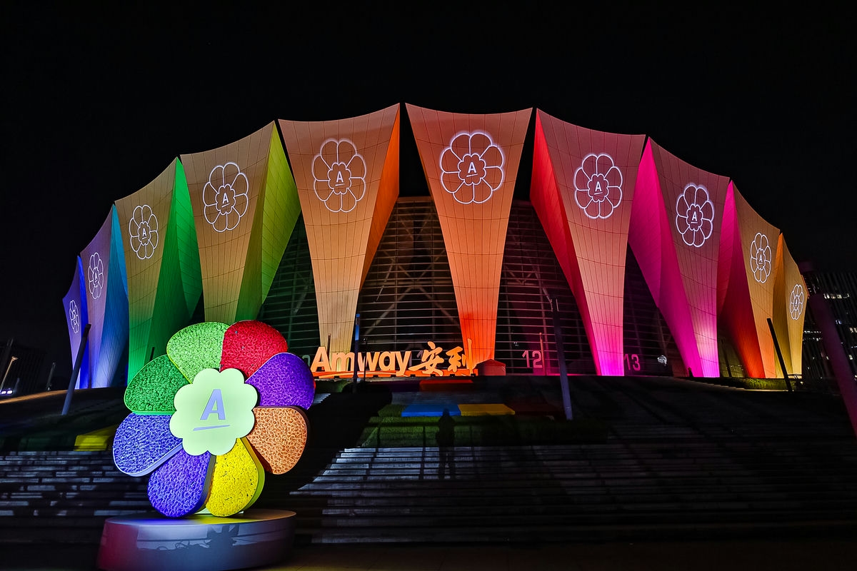 夜幕下的安利活动主场馆外立面铺满渐变彩虹色灯光，投射有安利专属花形标识，场馆入口处醒目展示“Amway安利”字样，台阶前放置着多彩的安利花形立体装置，整体灯光璀璨绚烂，氛围热烈隆重，尽显品牌活力与活动的盛大感。