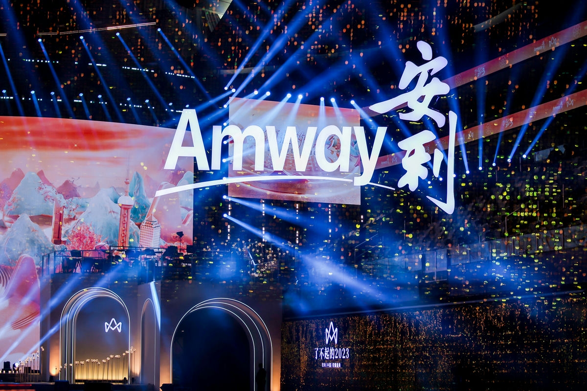 画面呈现安利品牌活动主舞台，醒目的“Amway”“安利”标识悬浮于舞台上方，蓝色光束交织，背景大屏融合国风山水与城市地标元素，舞台下方标注活动主题“了不起的2023”，场内星光点点，整体氛围恢弘热烈，尽显活动的盛大质感。