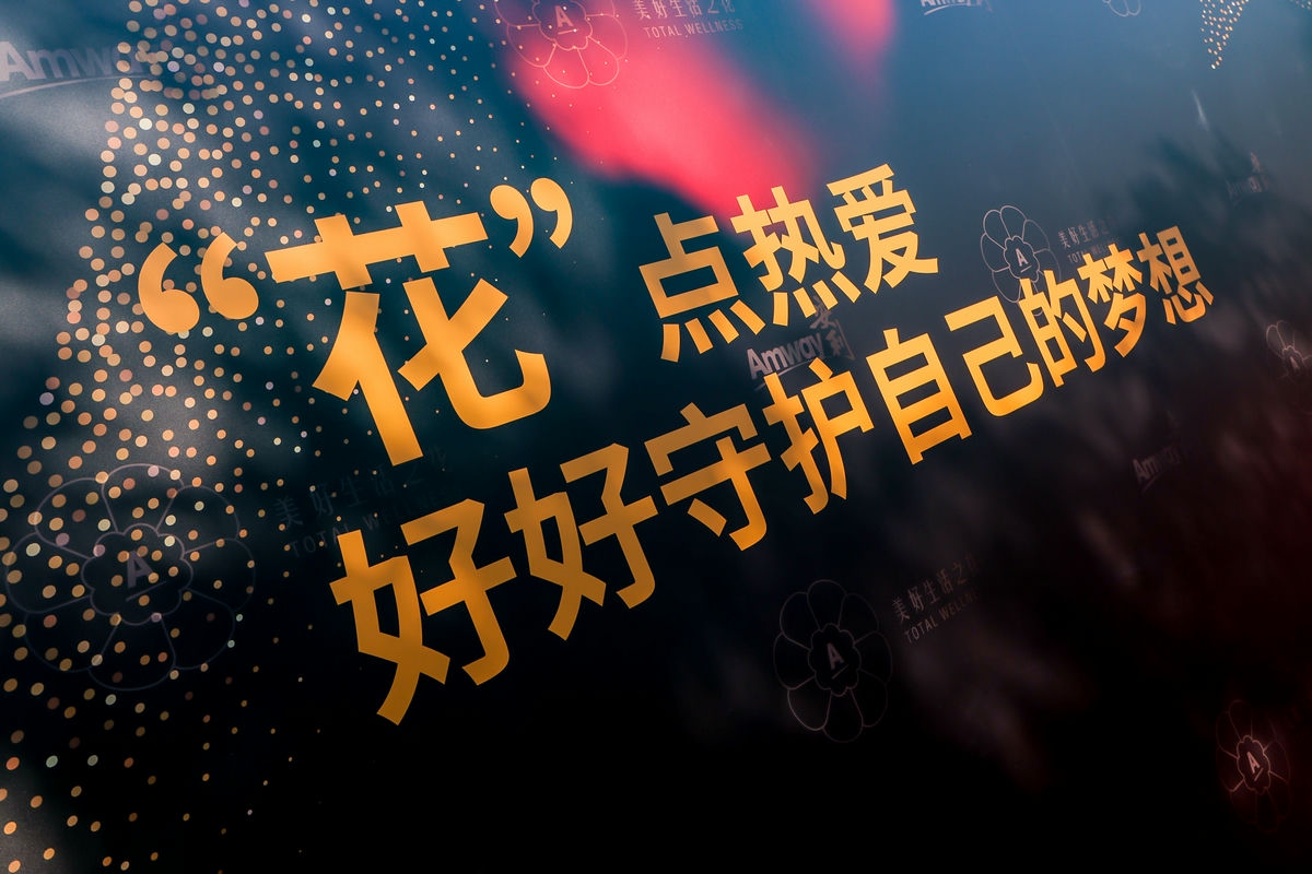 画面为安利活动宣传背景特写，深色底缀有闪亮点阵、安利品牌标识及“美好生活之心 TOTAL WELLNESS”字样，暖金色醒目文字写着“‘花’点热爱 好好守护自己的梦想”，整体氛围温暖积极，传递出安利鼓励大家奔赴热爱、逐梦前行的正向理念。