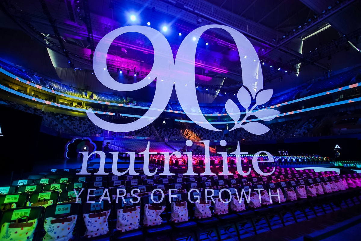 现场呈现醒目的“90”“nutrilite”“YEARS OF GROWTH”标识，开阔的大型场馆内坐席整齐排布，每座都放置了定制纪念袋，紫蓝炫彩灯光烘托出盛大热烈的氛围，尽显纽崔莱90周年庆典的隆重质感与品牌活力。