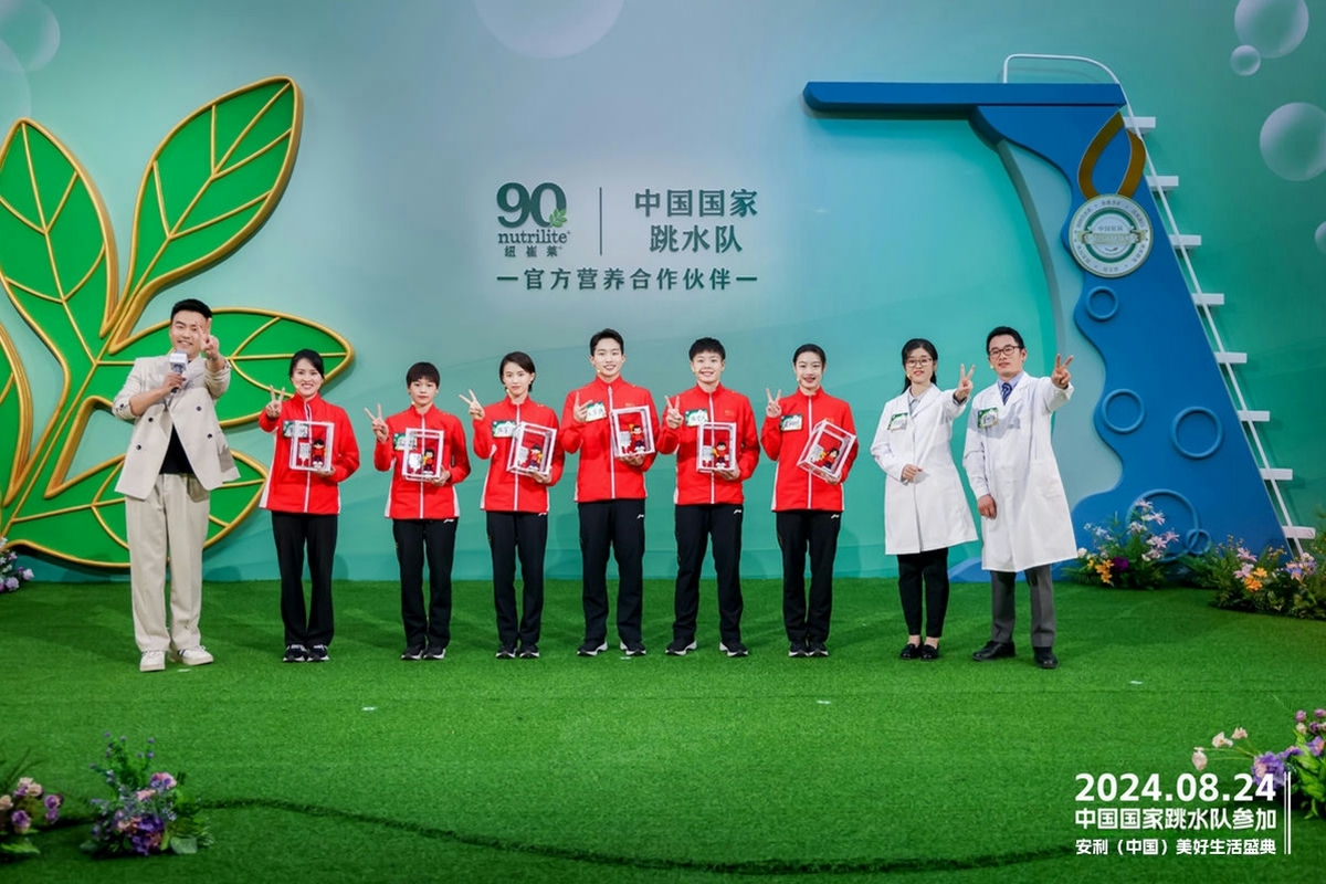 2024年8月24日安利（中国）美好生活盛典现场，主持人、中国国家跳水队成员与安利研发人员共同合影。背景标注纽崔莱90周年、纽崔莱为中国国家跳水队官方营养合作伙伴，众人手持纪念礼品面带笑容比出手势，氛围轻松热烈，尽显积极健康的活力风貌。