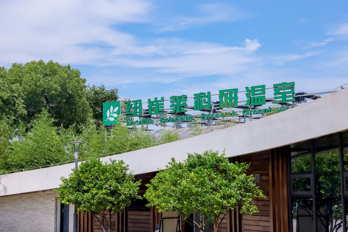 画面呈现晴朗碧空下的纽崔莱科研温室外观，建筑顶部“纽崔莱科研温室”与对应英文标识醒目清晰，周边绿植葱郁繁茂，整体氛围清新明快，彰显纽崔莱在自然营养领域的科研硬核实力。