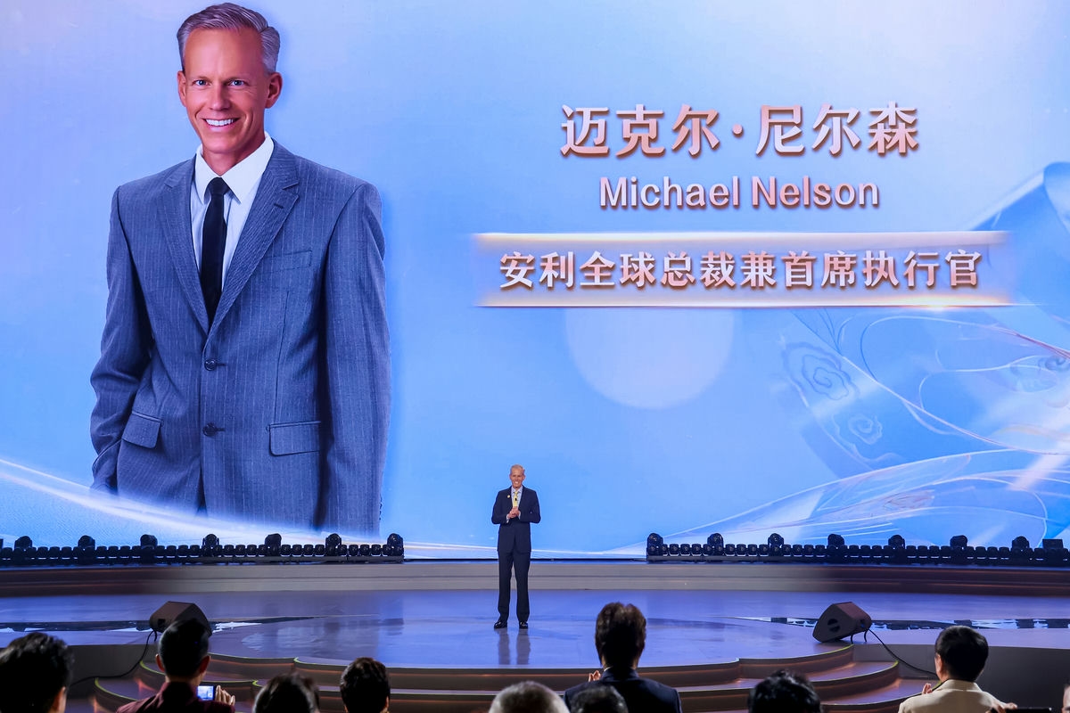活动现场，安利全球总裁兼首席执行官迈克尔·尼尔森（Michael Nelson）站在舞台中央发言，身后巨幅大屏同步展示他的个人形象与职务信息，台下参会人员认真聆听，现场氛围庄重热烈，尽显安利品牌活动的高规格与正向风貌。