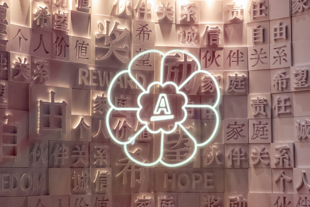 画面为安利活动文化展示墙，背景立体字牌镌刻着诚信、希望、责任、伙伴关系、个人价值、家庭、奖励、HOPE、REWARD等品牌核心价值关键词，墙中央是内嵌品牌A标识的花形霓虹造型，整体氛围温暖厚重，彰显安利正向文化内核。