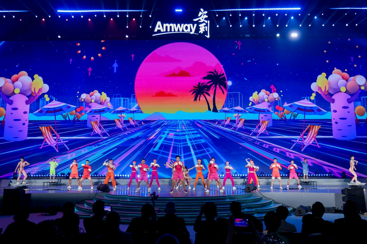 舞台上方醒目展示“Amway安利”品牌标识，背景大屏是落日椰树的炫彩动感度假风画面，身着亮色系运动服的表演者在台上整齐演绎健身操，动作饱满富有活力，台下观众认真观演，现场氛围热烈鲜活，尽显安利积极健康的品牌调性。