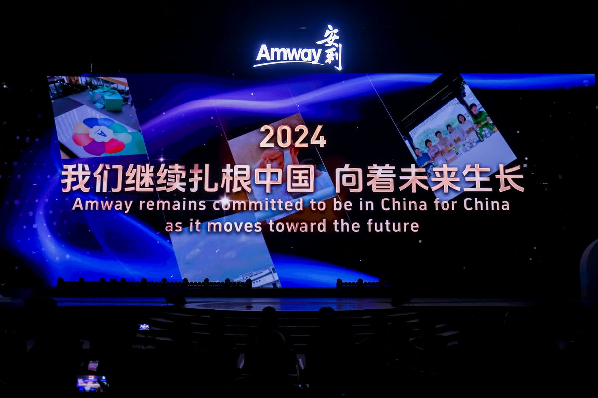 活动现场顶部醒目点亮Amway安利品牌标识，主屏幕展示2024“我们继续扎根中国 向着未来生长”的主题及对应英文标语，搭配品牌相关场景实拍素材，蓝紫色流光背景烘托出昂扬庄重的氛围，传递出安利深耕中国市场、拥抱未来发展的坚定决心。