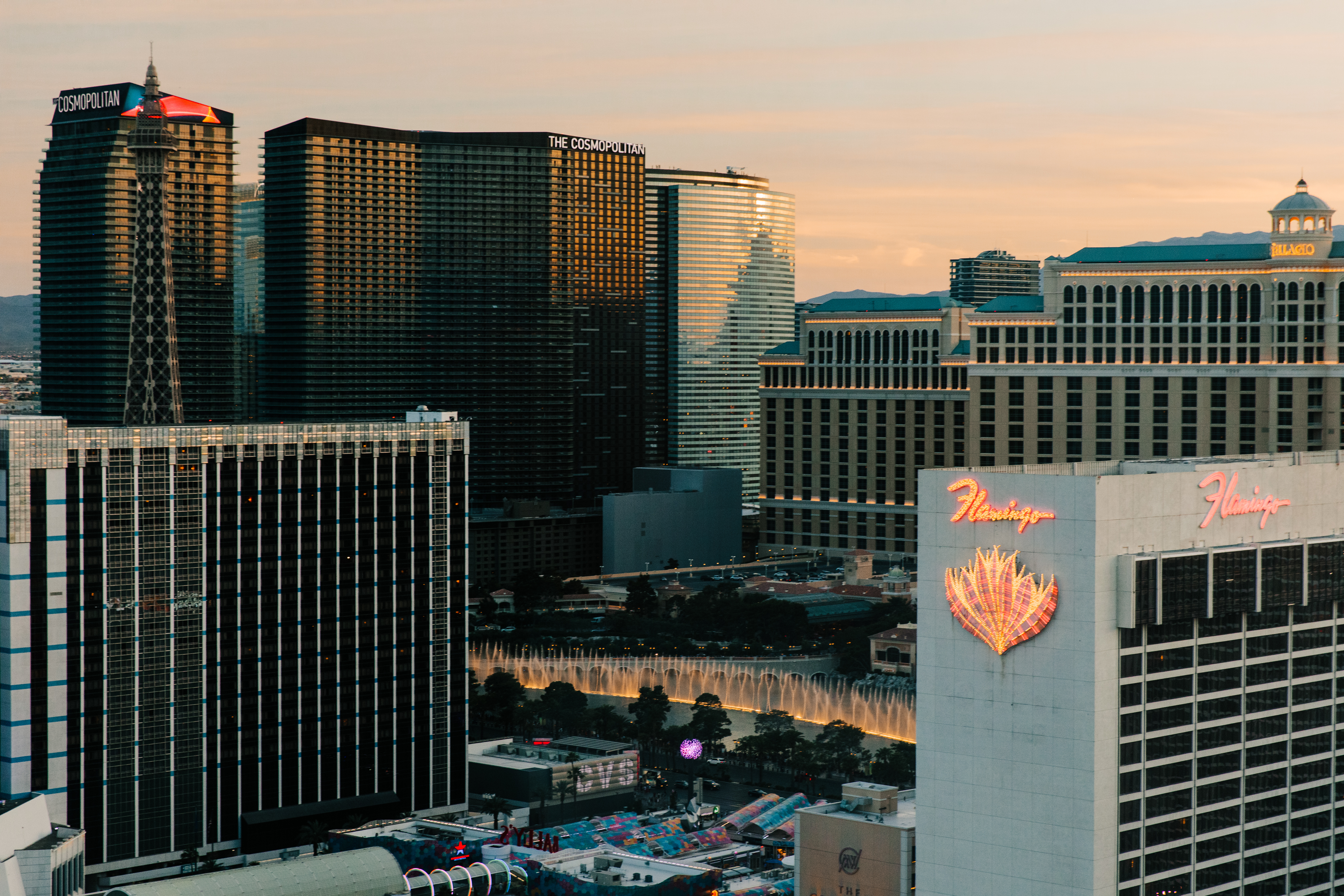 黄昏暖光铺洒在拉斯维加斯核心城区，画面可见“COSMOPOLITAN”“Flamingo”等地标标识，百乐宫喷泉灵动涌动，摩天楼宇错落林立，整体氛围开阔璀璨，尽显国际都市活力，适配安利海外高端会务活动选址需求。