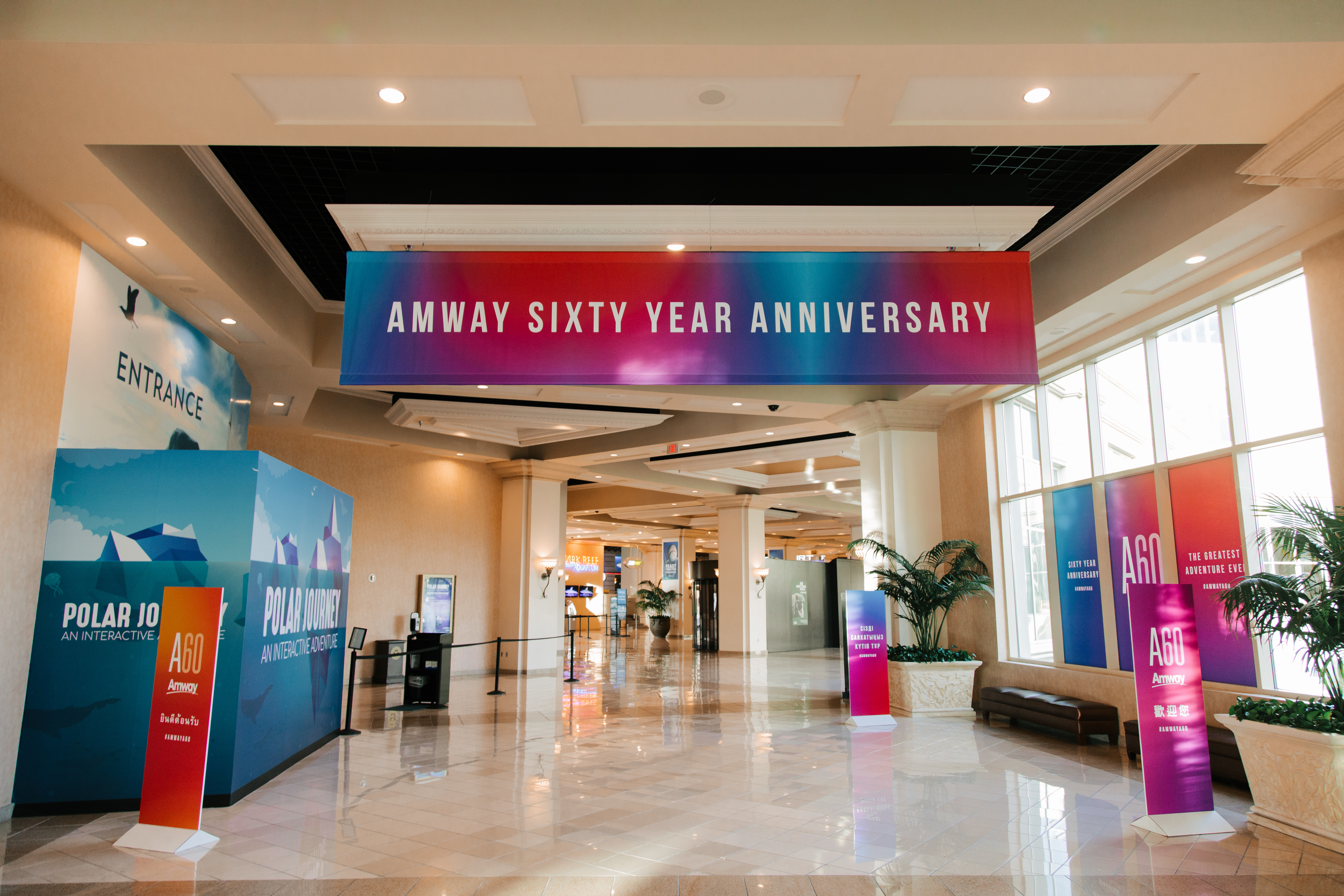 画面为安利60周年活动会场入口，上方悬挂“AMWAY SIXTY YEAR ANNIVERSARY”主题横幅，大厅宽敞明亮，两侧设有印有“A60”“欢迎您”字样的主题立牌、“POLAR JOURNEY”互动区装置，布置规整大气，氛围隆重热烈，尽显活动盛大质感。