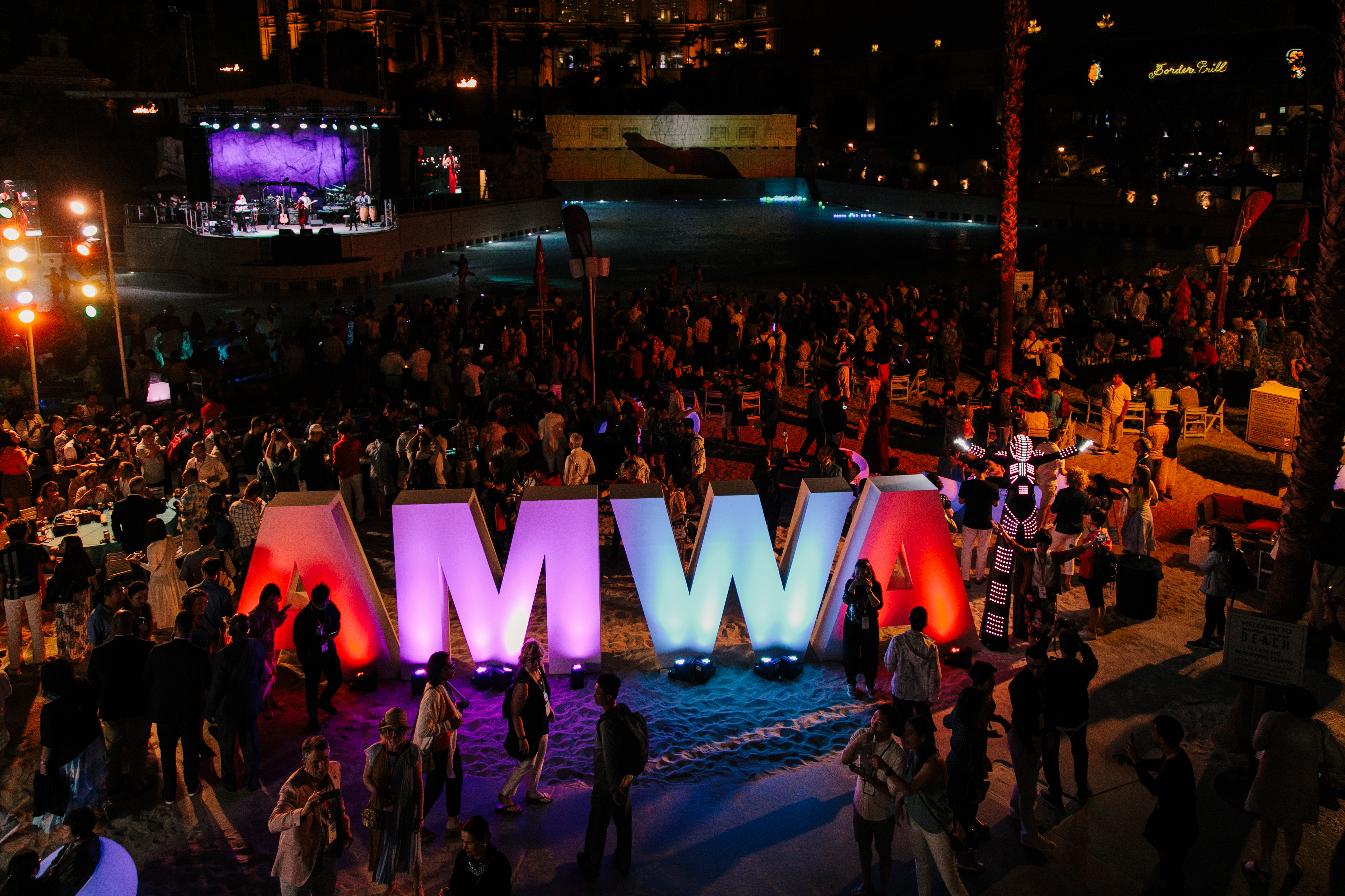 夜色下的沙滩活动场地中，巨型渐变发光“AMWA”立体标识十分醒目，远处舞台灯光绚烂，众多参会伙伴齐聚于此，特色演艺装置穿插在人群间，现场氛围热烈欢快，充分彰显安利活动的活力与超高凝聚力。