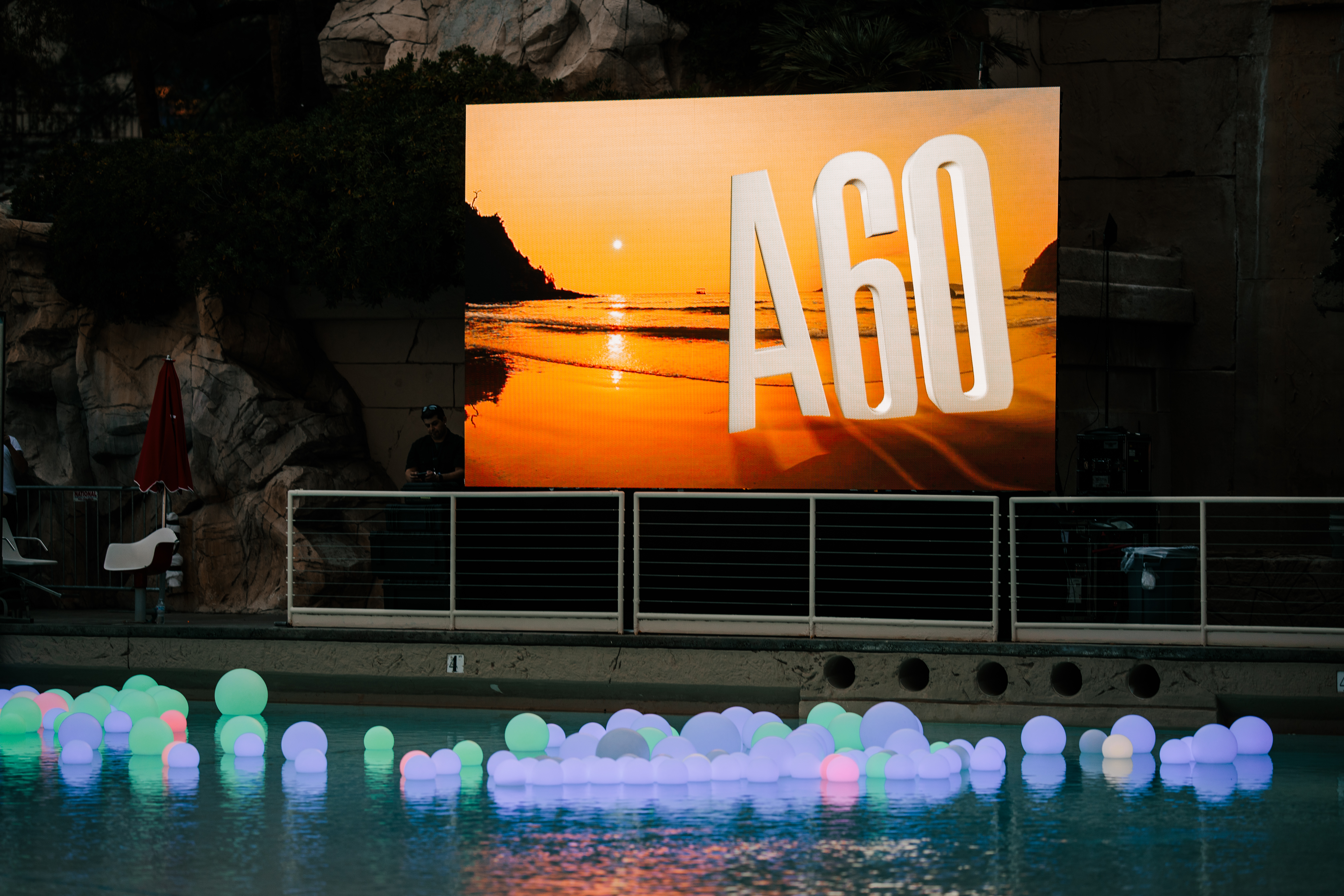 画面为安利A60周年活动户外场地，大屏展示醒目“A60”字样，搭配暖调海滨落日背景，前方泳池中漂浮着多色发光圆球，光影在水面柔化倒映，整体氛围温馨璀璨，尽显60周年庆祝活动的用心与活力。